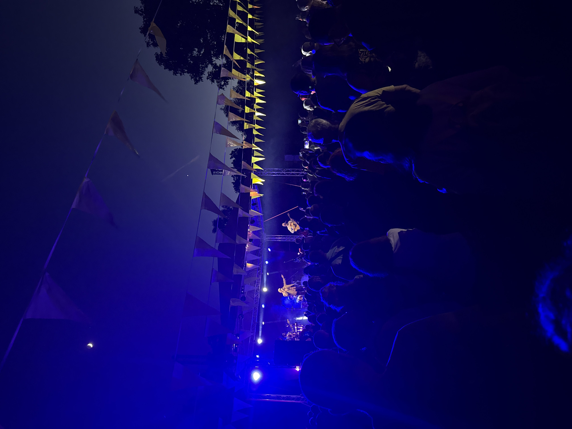 An outdoor concert is taking place at night, illuminated by bright blue stage lights. A band is performing on a raised stage, with several musicians visible playing instruments and singing. The audience, silhouetted in the foreground, stands or sits watching the performance. Festive triangular flags are strung across the sky above the stage and audience. The crescent moon and a faint star are visible in the dark blue twilight sky. The location is identified as Stara Zagora, Bulgaria.