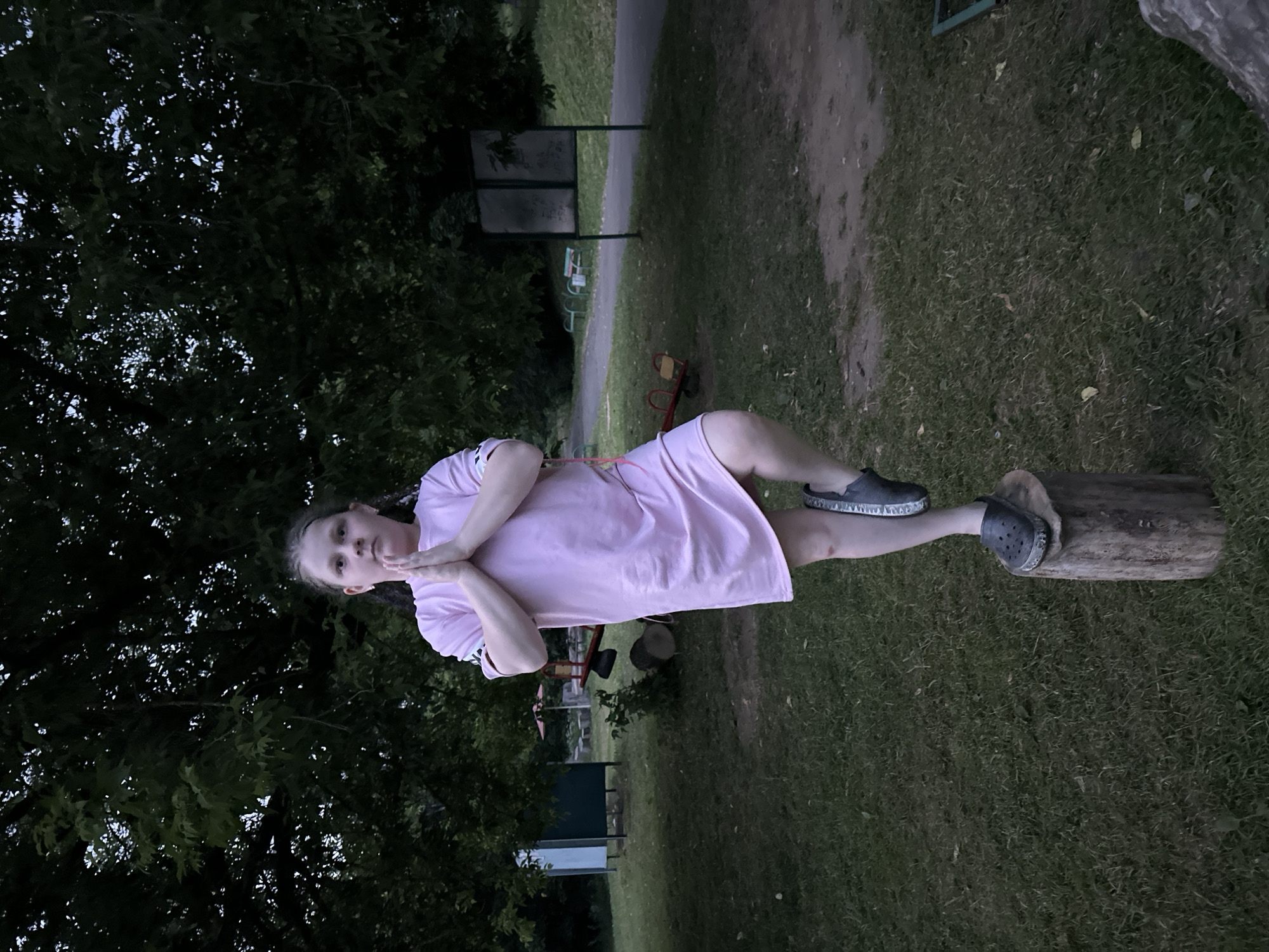 A young girl stands on a tree stump in Sokolovo, Russia, performing a yoga pose with her hands pressed together in front of her chest and her right leg bent at the knee. She is wearing a pink dress and grey clogs. The background features a grassy area with trees, a playground with a seesaw, and a path. The lighting suggests it is evening or dusk.