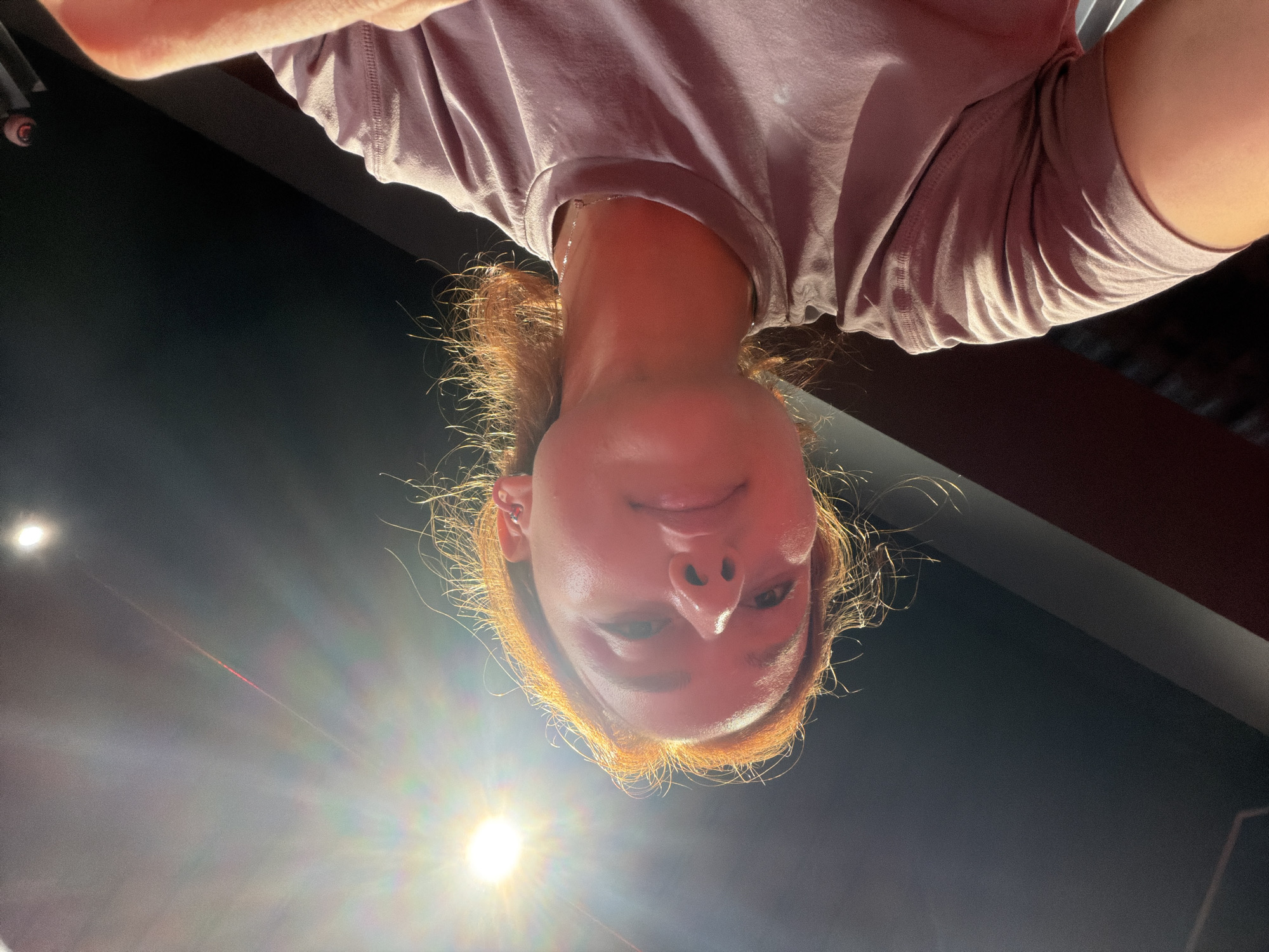 The image shows a low-angle, close-up, upward-facing selfie of a young woman with reddish-blonde hair. She is wearing a light pink t-shirt. The light source, a bright spotlight with visible lens flare, is positioned to the left and above her head, casting strong highlights on her face and hair. The background is dark, with a diagonal line suggesting a wall or ceiling structure. The woman's face is turned slightly to her left, and she is smiling faintly. Her right ear is visible, adorned with a small hoop earring. The overall lighting creates a dramatic effect, with sharp contrasts between light and shadow. There are no discernible elements indicating a specific location within the city of Strasbourg, France.