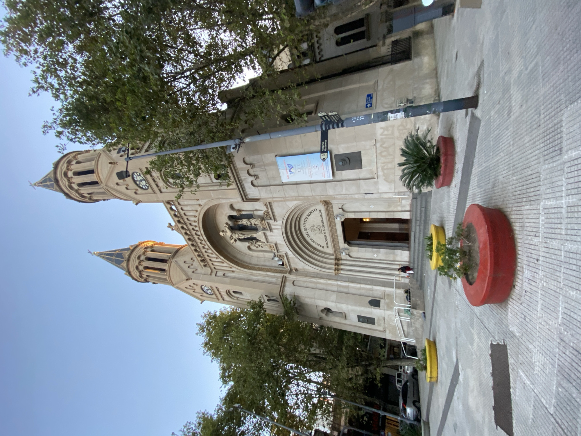 The image shows a daytime, exterior view of the Basilica del Espiritu Santo, a Roman Catholic church located in the Palermo neighborhood of Buenos Aires, Argentina. The church is constructed in a European style, featuring a stone façade, two prominent towers, and an arched entrance adorned with religious statuary. A street lamp is visible in front of the church, and a sign indicates the direction of "Hospital Gutierrez." Trees surround the building, obscuring some of the architectural details. In front of the church's entrance, planters in red and yellow can be seen. Sidewalk stones lead to the church entrance. A few figures, possibly individuals entering or exiting the basilica, are seen in the entrance. A few parked cars can be seen. The sky is blue.