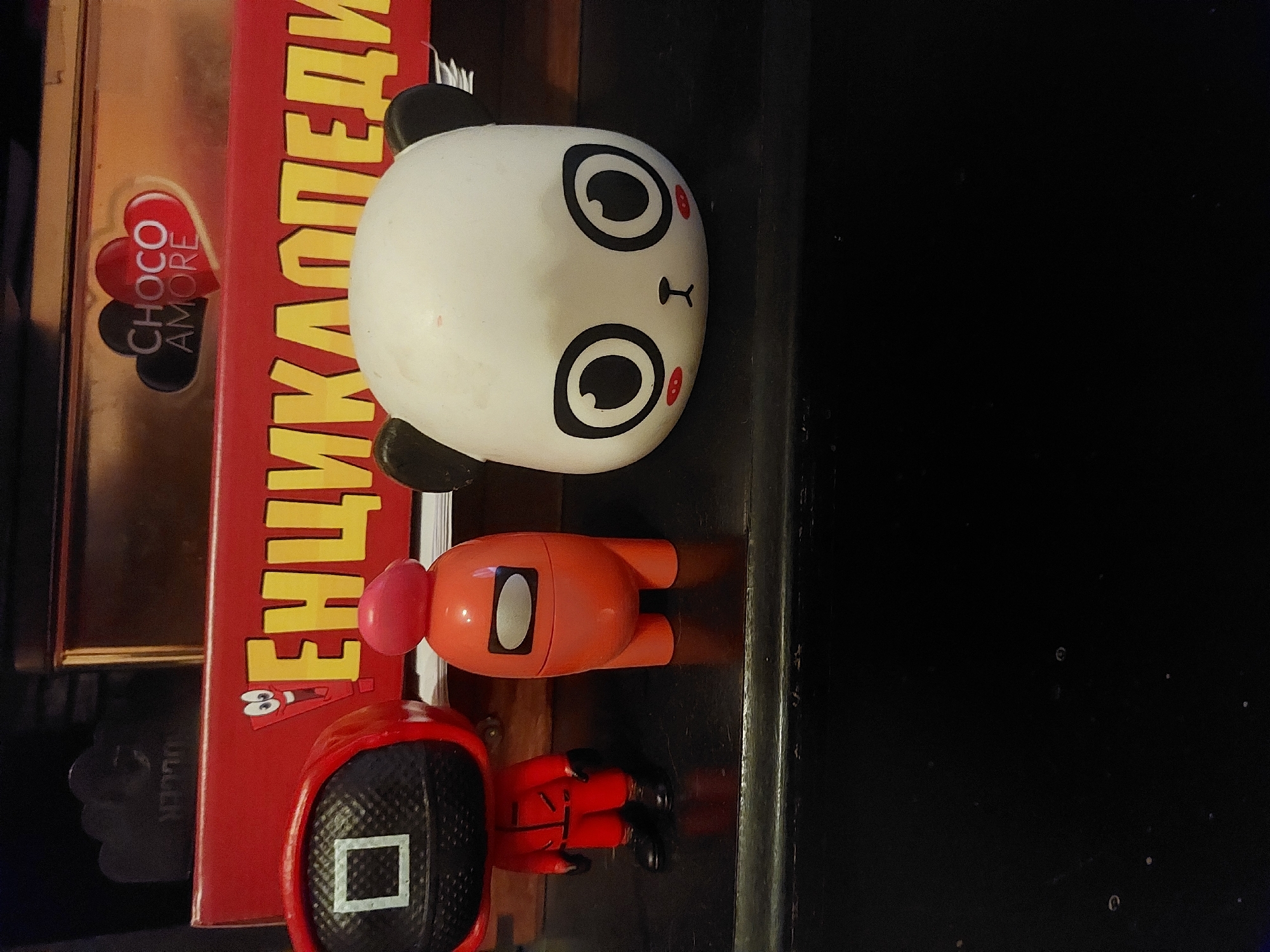 The image shows a collection of items on a surface. From left to right there is a box labeled "CHOCO AMORE," a red book with text written in Cyrillic, a panda-shaped object, an orange "Among Us" figure, and a "Squid Game" figure. The surface appears to be dark.
There is no evidence to indicate the city of Tsarevo, Bulgaria.