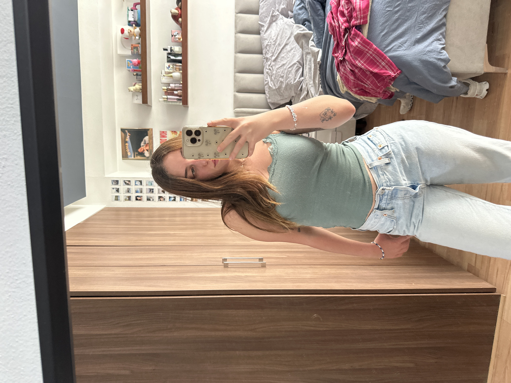 A woman is taking a mirror selfie indoors. She is wearing a green tank top and light blue jeans. Behind her is a bed with blue and grey bedding, and a shelf with various small items. There is a wooden dresser in front of the mirror. The location is likely a private residence. The city, Huixquilucan de Degollado, Mexico, is assumed as the person asked for it, but not readily apparent in the picture itself.