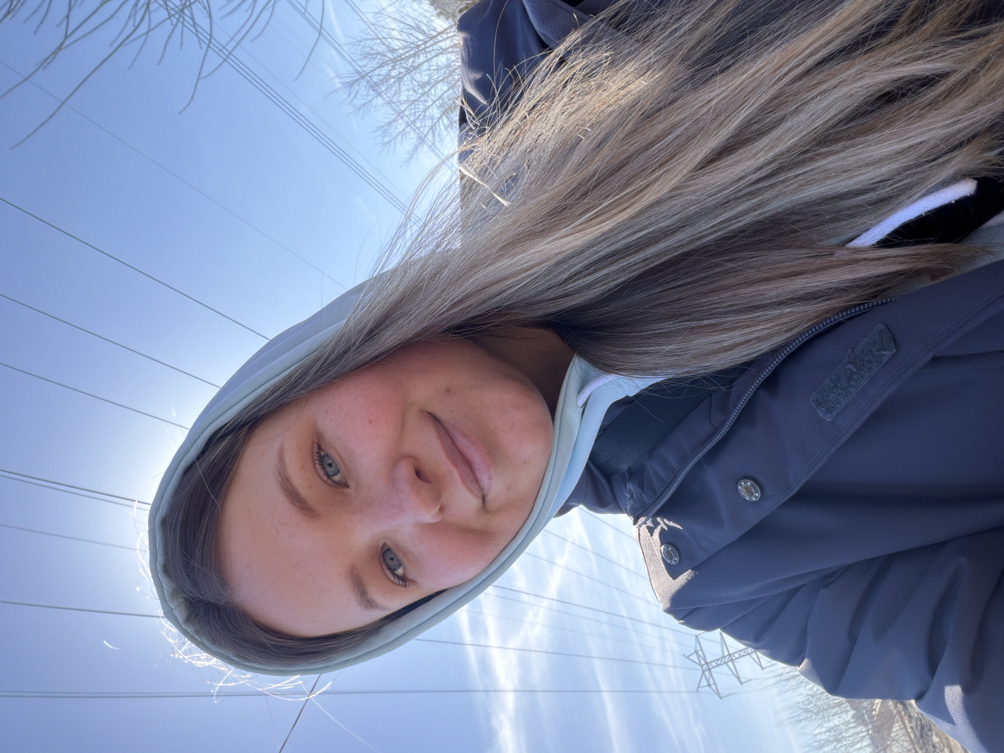 A photograph depicts a young woman in a hooded jacket with long, light-brown hair. The photo is taken from a low angle, looking up at the subject. The background shows a clear, light-blue sky with power lines visible above. Bare tree branches are present in the corners of the image. The location is indicated to be Chaikovsky, Russia.