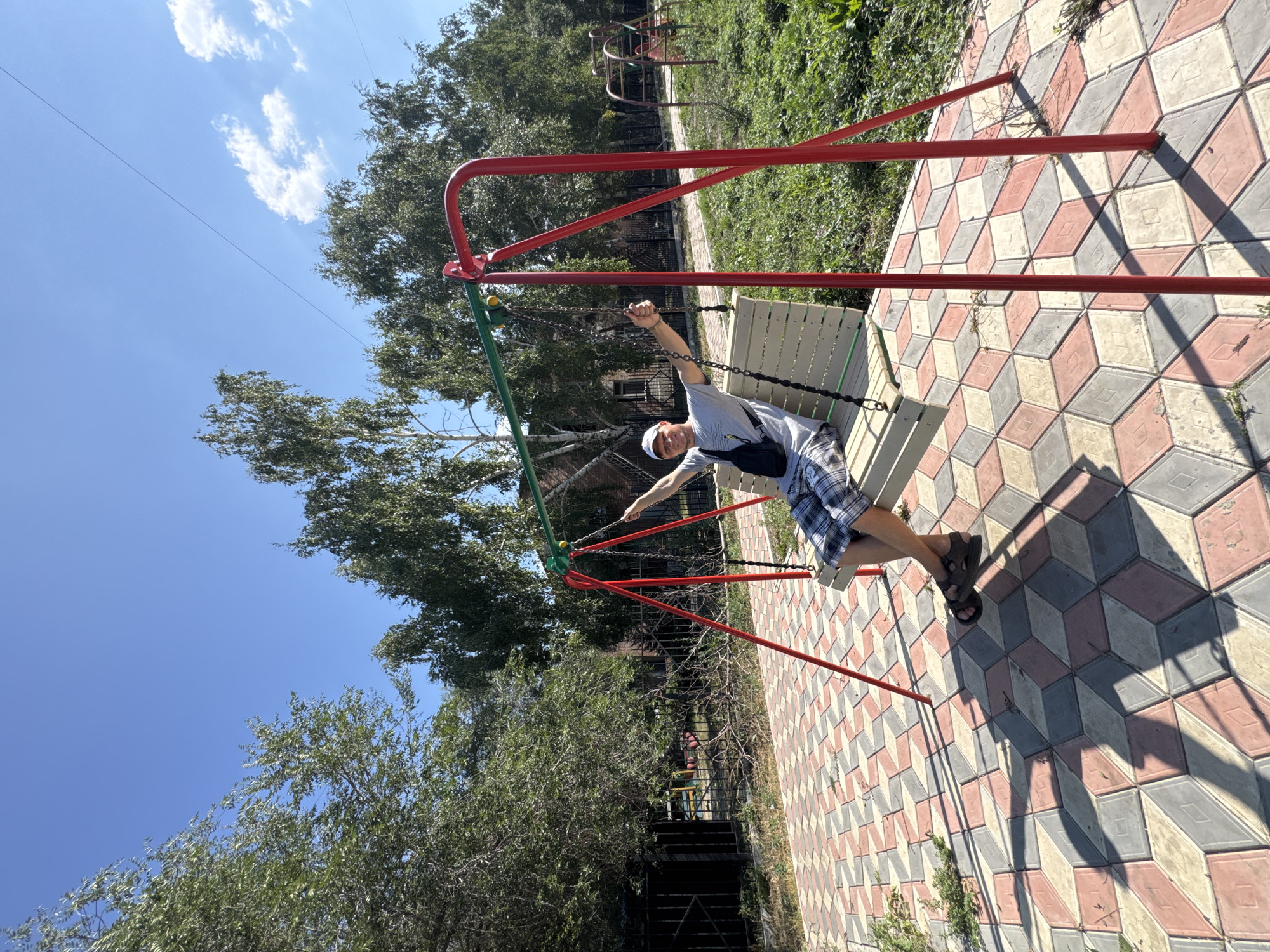 A man sits on a swing in a park. He is wearing a grey t-shirt, plaid shorts, sandals, and a white cap. He has a black bag slung across his chest. The swing set has red metal supports and a green crossbar. The man is holding onto the chains of the swing with his hands raised. The ground beneath the swing set is paved with colorful interlocking bricks. In the background, there are trees, a fence, and playground equipment. The sky is blue with some white clouds. The overall setting appears to be a children's playground in a city.