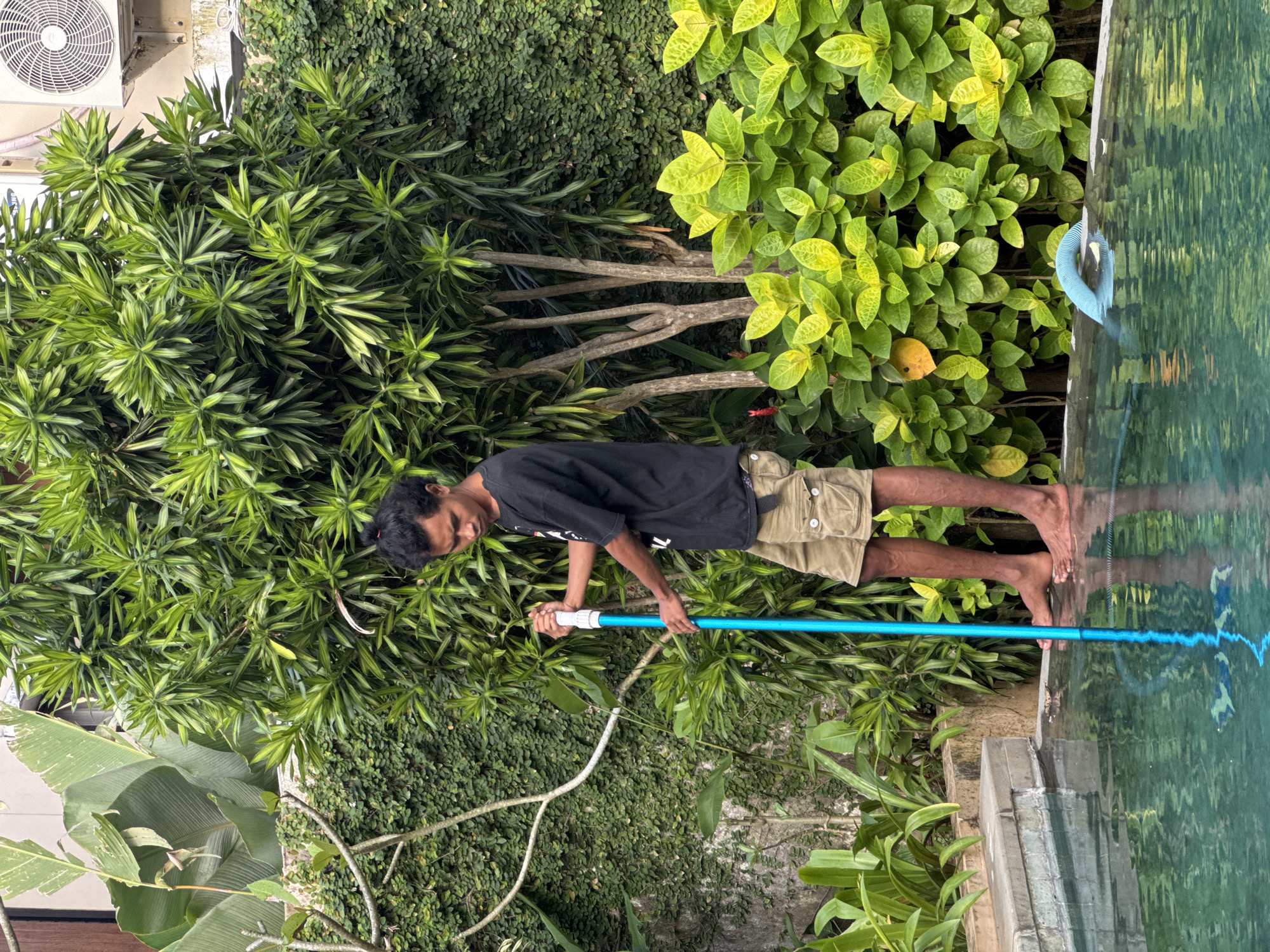 A person is standing beside a pool in an outdoor setting in Kuta Selatan, Indonesia. The person, who appears to be male, is wearing a dark t-shirt and khaki shorts. He is holding a long blue pole and appears to be cleaning or maintaining the pool. His feet are near the edge of the pool. Lush greenery surrounds the scene, including various types of plants and vines growing on the wall and along the poolside. An air conditioning unit is mounted on a wall above the plants. The pool water reflects the surrounding vegetation.