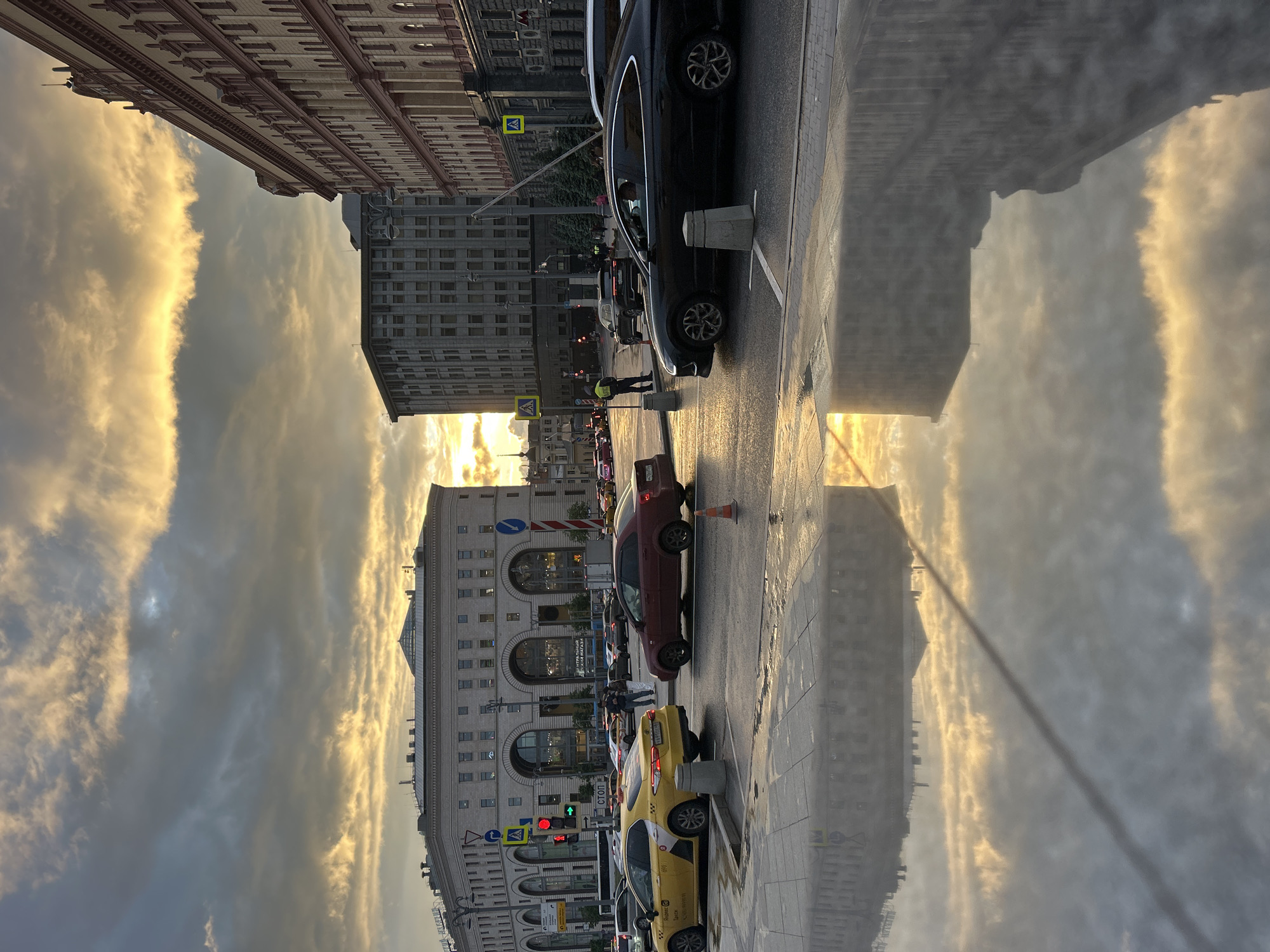 The image shows a street scene in an unknown city. Several buildings are visible, including one with arches. Multiple cars are parked along the side of the road, including a black SUV, a maroon sedan, and yellow taxis. A traffic light is red. A person in a yellow vest stands near the curb. The sky is cloudy with golden light. The wet street reflects the buildings and sky.