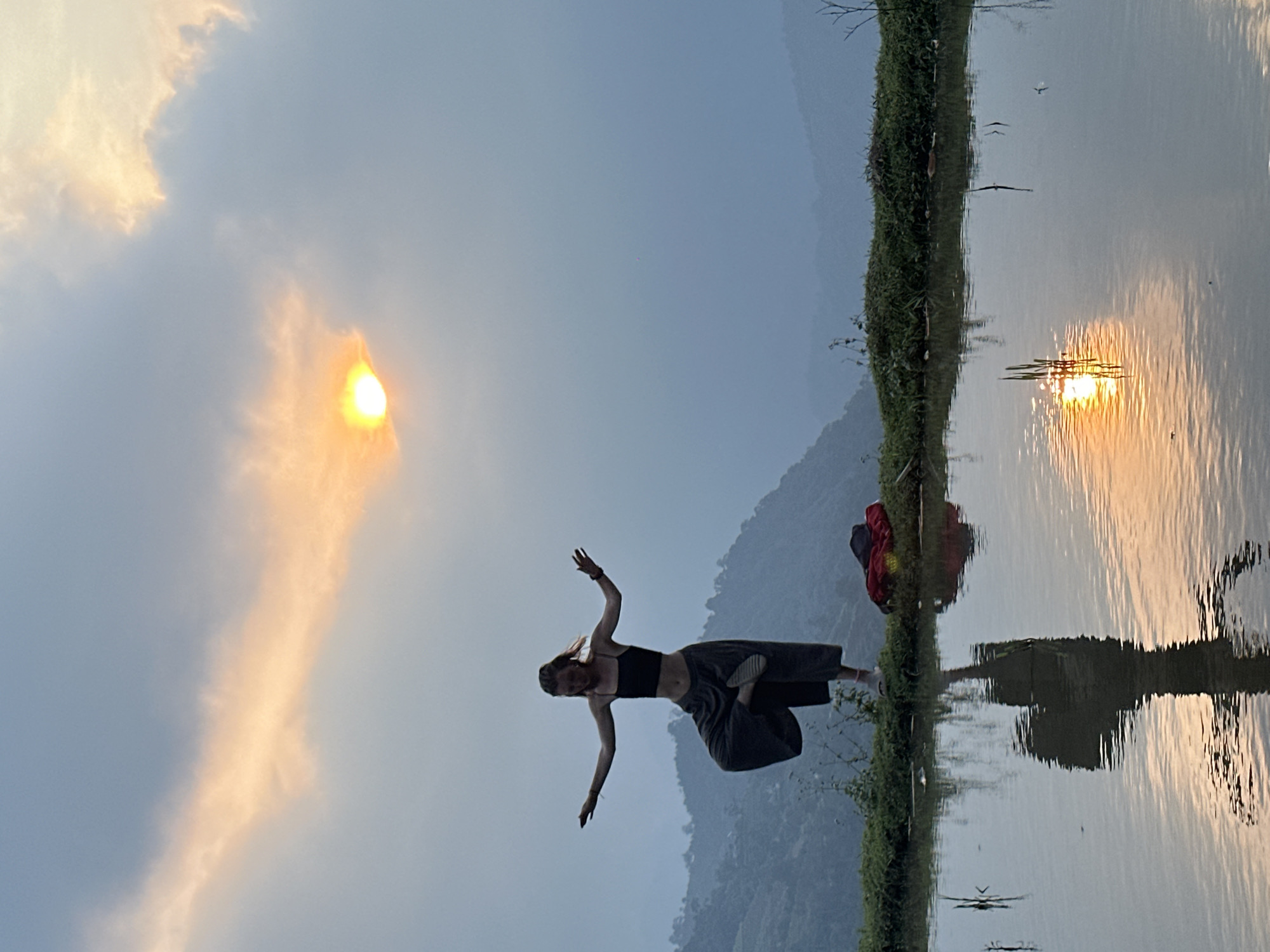 The image depicts a serene outdoor scene during golden hour, likely sunset or sunrise, in Nậm Dịch Commune, Vietnam. A woman is positioned on a narrow, verdant strip of land bordering a calm body of water, possibly a lake or large pond. She is shown from approximately the waist up, wearing a dark crop top and dark, wide-leg trousers. Her light-colored hair is visible, and her arms are outstretched with one leg bent, suggesting she is mid-pose, perhaps engaging in yoga or dance.
Behind the woman, a red and dark-colored bag or backpack rests on the grassy bank. The background reveals a landscape of hazy, mist-shrouded mountains under a sky with scattered clouds. The sun, a bright, golden sphere, is visible in the upper part of the sky, casting a warm, glowing light that permeates the clouds. Its shimmering reflection is prominently displayed on the tranquil water surface, creating elongated streaks of light. The water also provides a distorted reflection of the mountains, the woman, and the bag. Small, dark shapes, possibly birds or insects, can be seen both in the sky and just above the water. The overall scene conveys a sense of tranquility and connection with nature. No visible text is present in the image.