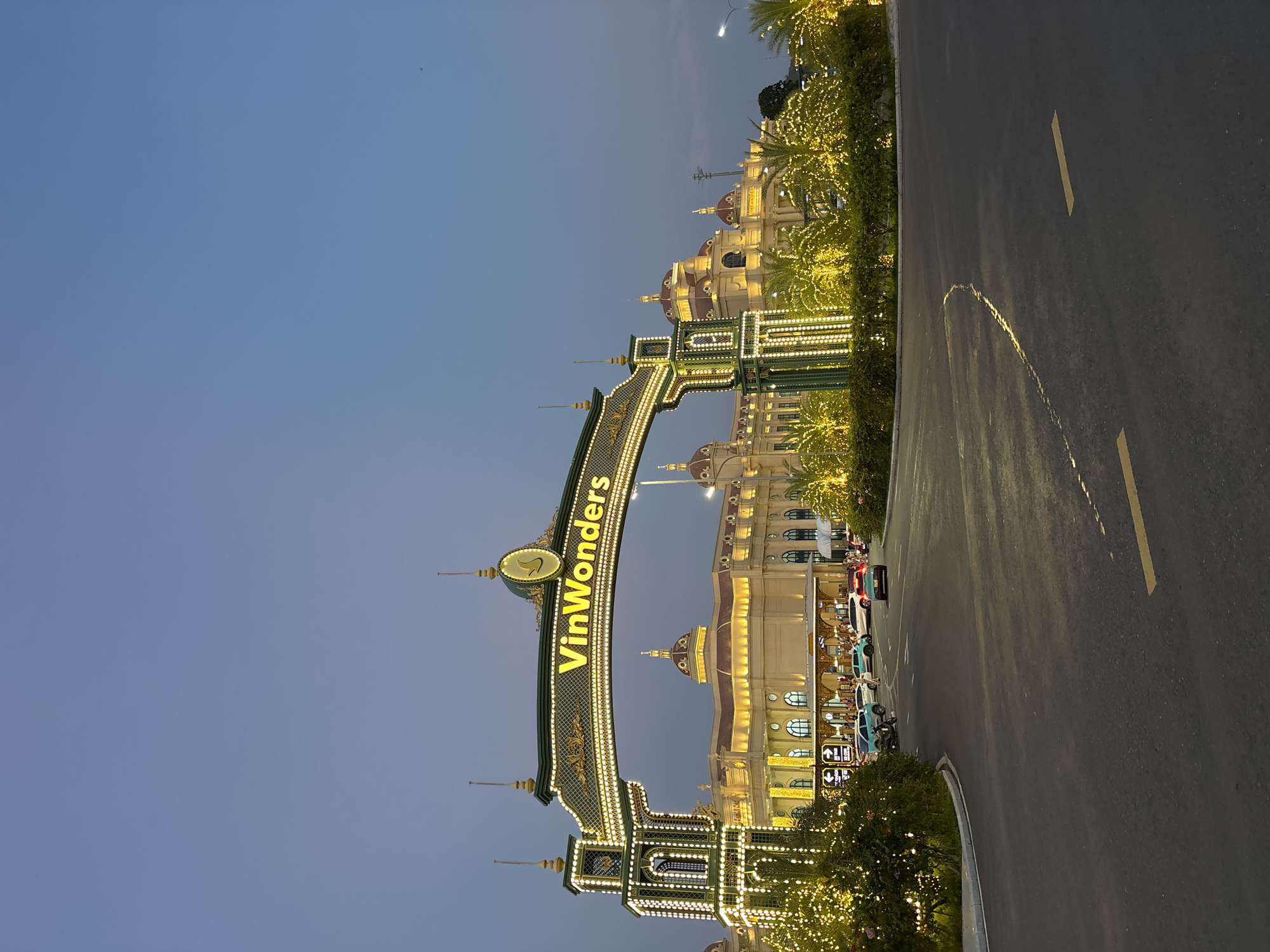 The image captures the illuminated entrance to "VinWonders" in Nha Trang Ward, Vietnam, during dusk or early evening. A grand, ornate archway, richly decorated with gold and green accents and numerous small, bright lights, spans across the scene. The name "VinWonders" is prominently displayed in large, glowing yellow letters on the archway's main beam.
Behind the archway, several large, stately buildings with classical European architectural styles are visible, featuring multiple domed roofs and arched windows. These structures are also brightly lit, emphasizing their grandeur against the darkening sky. Along the roadside and framing the archway, dense foliage and palm trees are adorned with countless twinkling lights, contributing to a festive atmosphere.
An asphalt road occupies the right side of the frame, showing yellow lane markings and the faint reflection of lights. Several vehicles, including cars and motorbikes, are parked or driving near the buildings. Small, lit directional signs can be seen near the parked cars. The sky above is a clear, deep blue, indicating the transition from day to night. A tiny, faint speck is visible in the upper portion of the sky. The scene conveys a sense of arrival at a vibrant and elaborate entertainment destination.
