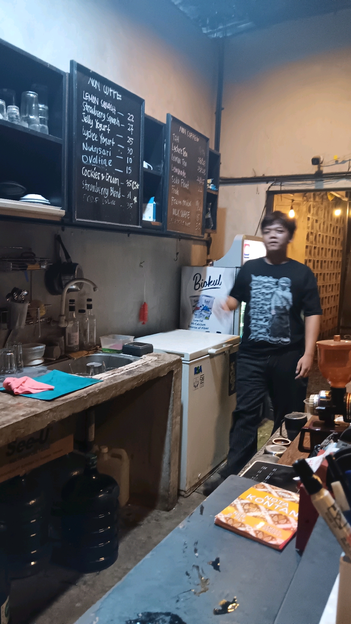 This media file depicts a young man standing in what appears to be a small cafe or food stall, likely located in South Jakarta, Indonesia. The setting is dimly lit, with a rustic and somewhat cluttered aesthetic, suggesting an indoor establishment.
The primary subject is the young man, who is casually dressed in a black t-shirt with a graphic print and dark pants. He is looking towards the viewer and appears to be in motion, possibly talking or gesturing.
Objects prominently featured include a kitchen counter with a sink, bottles, and a pink cloth, alongside a refrigerator branded with "Biokul." There are also menu boards displayed on the wall behind the counter, listing various drinks. One menu board has the heading "NON OFFE" and lists items like "LEMON SQUASH," "Jelly Yogurt," and "Cockies & Cream" with their prices. Another board reads "NAN COPPEM" and lists teas and milkshakes. Below the counter, several large water jugs are visible. On a table in the foreground, there is a small book titled "NOTA KONTAN," suggesting a point-of-sale or order-taking area. A coffee grinder is also partially visible on the right side of the frame.
The scene appears to be set during either evening or nighttime due to the lighting conditions. The overall atmosphere is casual and functional, indicative of a local business. The man's expression is neutral, and there are no strong emotional cues. The presence of the "Biokul" branding, a well-known Indonesian dairy product, and the Indonesian text on the menus reinforce the location context.