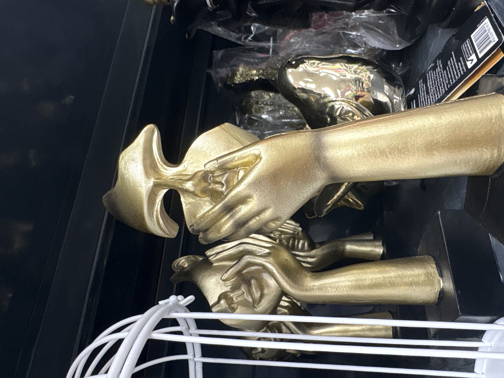 The image displays a close-up, angled view of several gold-colored decorative items, primarily stylized human hands and forearms, arranged on what appears to be a black shelf or in a storage unit. These objects have a matte gold finish, with some reflective surfaces, and show textures suggestive of casting or painting. Some hands are depicted as if cradling or holding other abstract gold forms.
In the background, additional gold-colored items are visible, some partially encased in clear plastic packaging, indicating they are new merchandise. A black product tag is partially visible in the upper right, displaying a barcode with the numbers "4 610397 810783" and small, unreadable text in Latin script below it. The setting implies an indoor retail environment, such as a shop or market in Chaikovsky, Russia, or a storage area for decorative goods.
In the lower foreground, a white wire basket or rack partially obstructs the view. The scene is static, featuring inanimate objects with no discernable activity or human presence. The lighting suggests an indoor setting, and no specific time of day or weather conditions are evident. No emotions are conveyed by the subjects.