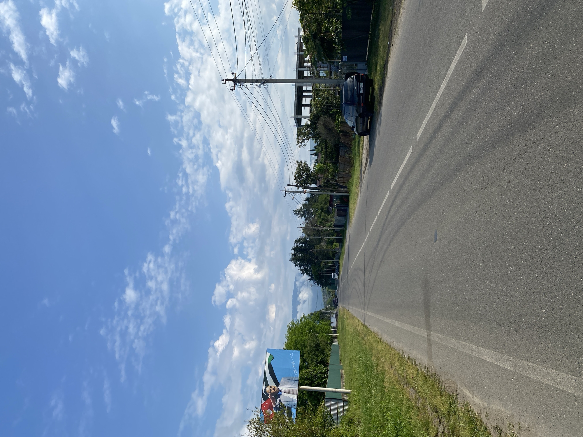 An eye-level, vertical shot captures a paved road stretching into the distance under a bright blue sky dotted with white, puffy clouds. On the left side of the road, a grassy verge borders a green metal fence, beyond which lush green trees line the landscape. A large billboard stands on the left, featuring a portrait of a man in a suit saluting, superimposed over national flags. Further down the road, a dark-colored SUV is parked on the right, near a construction site with a partially built concrete structure. Utility poles with numerous wires crisscross the sky above the road and the surrounding greenery. The road itself has white dashed and solid lines marking its lanes, and tire marks are visible on the asphalt. The scene conveys a sense of rural or suburban road infrastructure under a clear day.