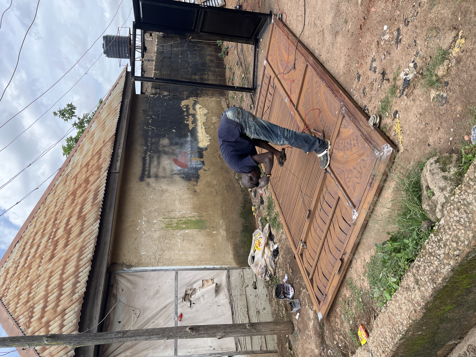 A person is working on a large, ornate metal gate. The gate is a reddish-brown color and has intricate patterns. The person, wearing a dark blue shirt and blue jeans, is bent over the gate, appearing to be welding or making repairs. They are wearing casual shoes.
The scene is outdoors, on a dirt ground covered with some debris, including plastic bags and scattered items. To the left of the gate is a dilapidated building with a rusty corrugated metal roof. The building's walls are rough and weathered, with some areas showing peeling paint and graffiti. Above the building, power lines crisscross against a cloudy sky. A black water tank is visible on a platform next to the building.
In the background, there is more of the building and a glimpse of vegetation. To the right of the gate, there is a rough concrete structure with moss growing on it, and more green plants. A partially opened black metal frame, possibly part of another gate or fence, is visible behind the person.
The lighting suggests it is daytime, but the cloudy sky may indicate overcast weather. The overall impression is one of a practical, outdoor workspace in a somewhat neglected urban or semi-urban environment. The activity is focused and appears to be a repair or construction task. There is a can of "GLOSS PAINT" visible on the ground near the gate.