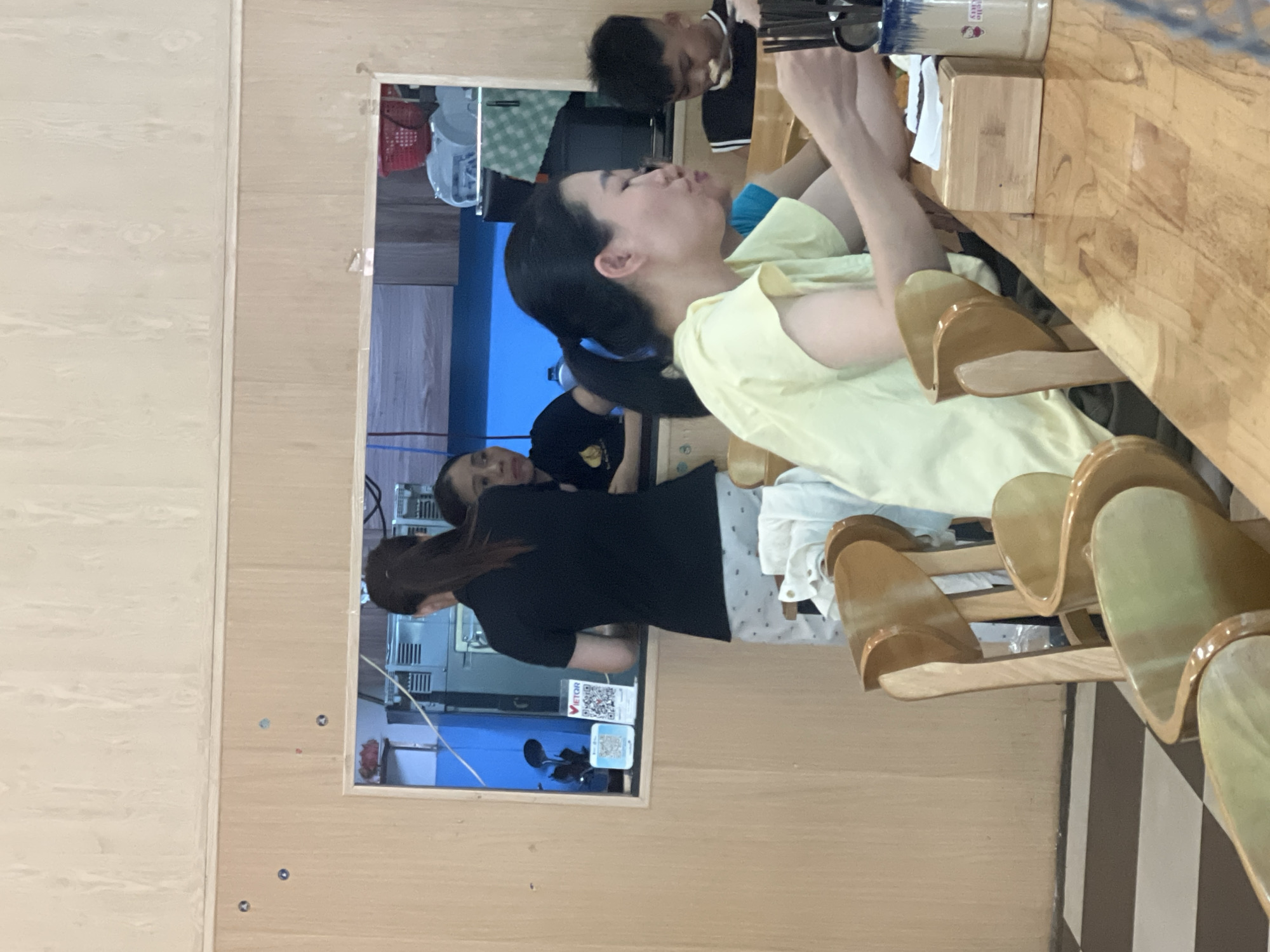 The image depicts the interior of a casual eatery in Đà Nẵng, Vietnam. In the foreground, a woman with her hair in a ponytail, wearing a pale yellow shirt, is seated at a wooden table, appearing to eat. Her mouth is slightly open, and she holds a utensil in her right hand. Beside her, another person, partially visible, is also seated. To the right, a young boy in a black and white striped shirt is eating from a bowl. In the background, through a rectangular opening, two women are visible behind a counter. One woman, wearing a black shirt and patterned shorts, stands with her back to the viewer. The other woman, in a black t-shirt, is looking towards the right. The environment appears to be functional, with wooden walls and furniture, and a checkered floor. QR codes are displayed on a shelf behind the counter.