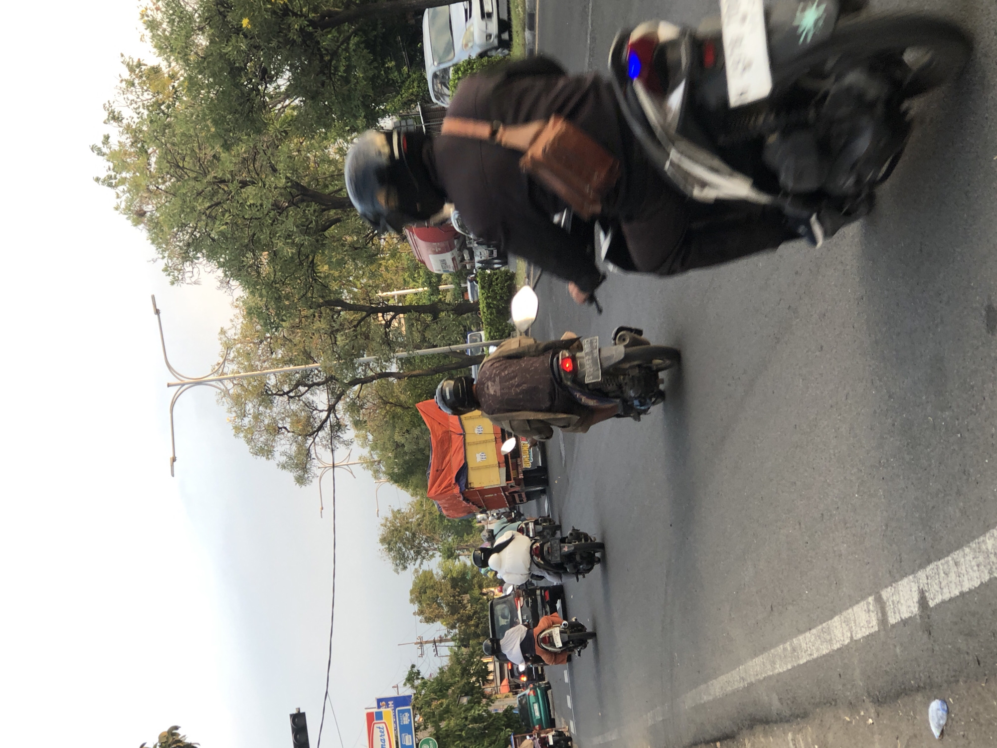 The image depicts a street scene in Kludan, Indonesia, with traffic, pedestrians, and a truck. In the foreground, a person wearing a black helmet and black clothing rides a motorcycle, with a brown satchel slung across their chest. To the left, another motorcyclist in a white outfit also travels on a motorcycle. Further back, several other motorcycles are visible, some carrying passengers. A large yellow truck with an orange tarp covering its load is in the middle of the road. A silver car is parked on the right side of the road. Trees line the street, and a traffic light is visible on the left side of the image. The sky is overcast.