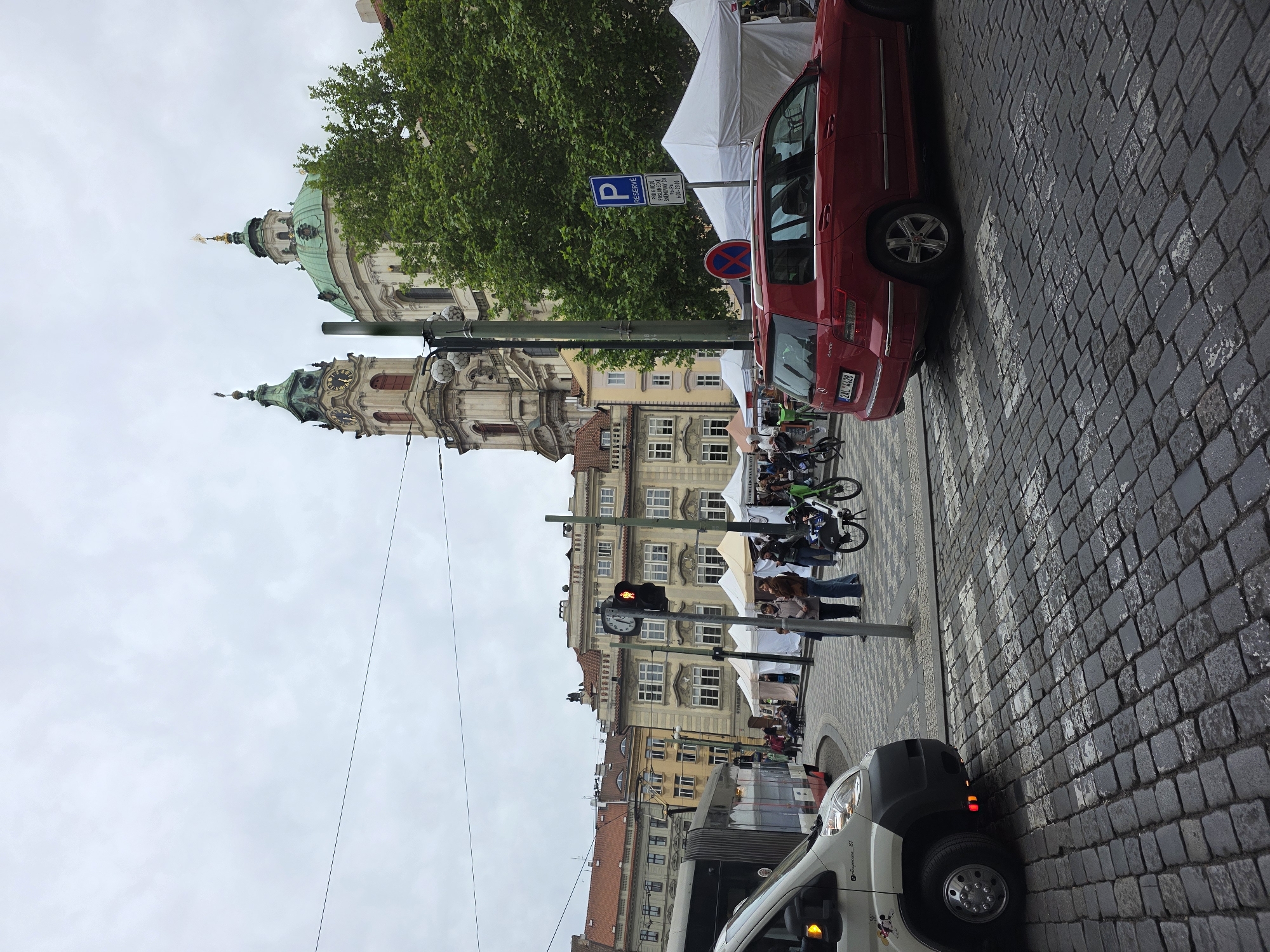 The image presents a street scene, likely in Prague, Czech Republic, featuring the iconic domes of St. Nicholas Church prominently in the background. The street is paved with cobblestones and includes a crosswalk. A red car and a white van are parked along the side of the road. A street lamp is visible with a built-in clock and traffic lights. There are a few people walking on the sidewalk. A blue parking sign is posted near the red car. White tents are seen closer to the building in the background, indicating a possible market or event. The sky is overcast.