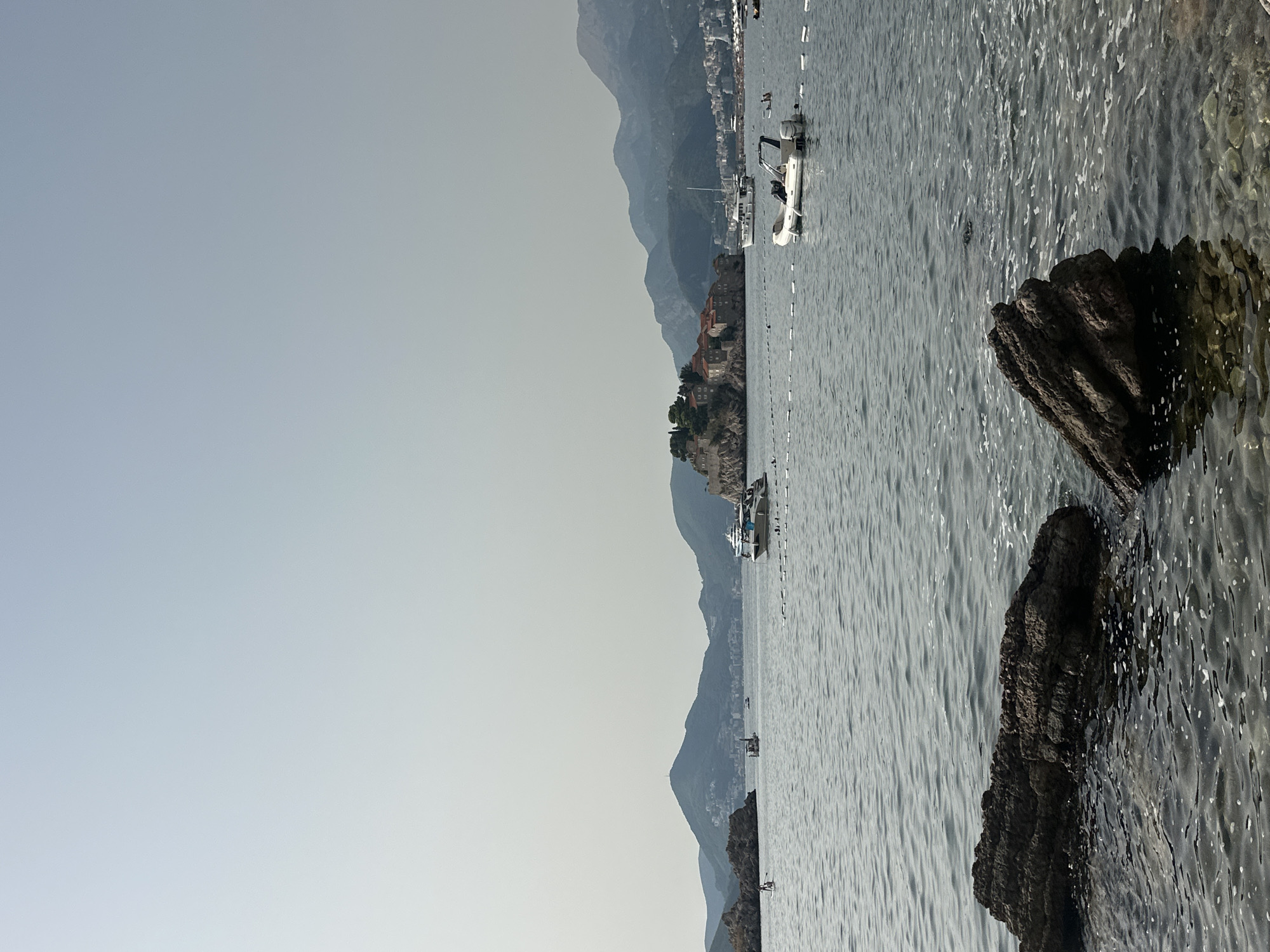 The image depicts a wide view of a calm, grayish-blue sea under a hazy, light blue sky. In the foreground, two large, dark, textured rocks are partially submerged in the shallow, clear water. The water surface is disturbed by gentle ripples and small white bubbles. In the middle ground, several boats are visible on the water. A prominent small island with red-roofed buildings and greenery, identified as Sveti Stefan, Montenegro, is situated in the distance, connected to the mainland by a narrow causeway. Hills and mountains form the backdrop, with some structures and trees visible on their slopes. There are several buoys marking off an area of the water. A few distant figures are scattered along the shoreline and in the water. The overall atmosphere is serene and coastal.