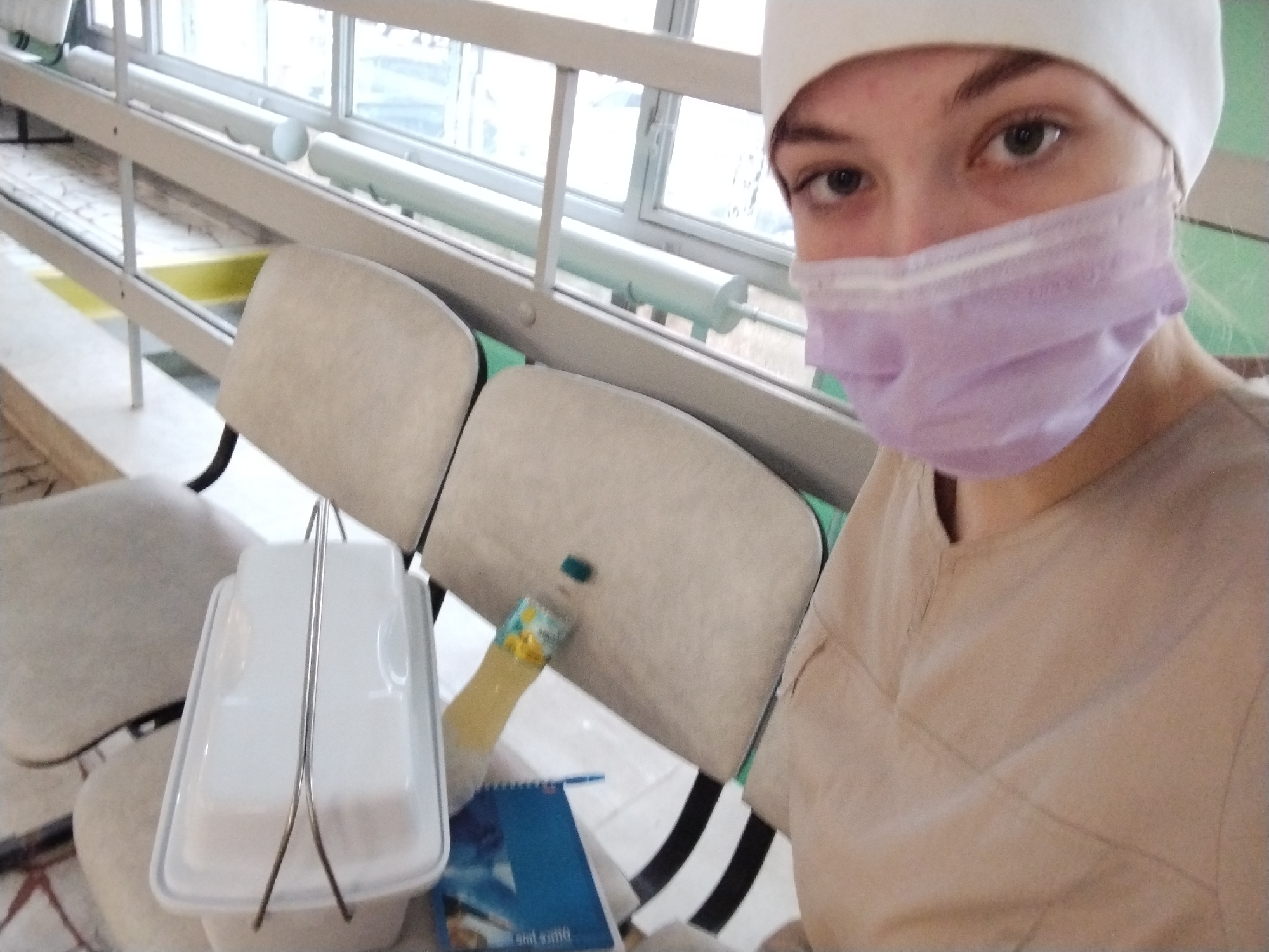 The image shows a person, likely female, taking a selfie indoors. She is wearing a white cap, a light purple disposable medical mask covering her nose and mouth, and a light beige scrub-like uniform top. Only her eyes are fully visible, looking directly at the camera with a neutral expression.
To her left, in the foreground, sits a white rectangular food container with a metal carrying handle, possibly a lunchbox. Behind this, several light-colored upholstered chairs are arranged in a row, facing away from the camera, suggesting a waiting area or common room. On one of the chairs or a surface nearby, there is a small plastic bottle with a yellow label and a green cap, and a blue notebook or book. Partially visible text on the blue item, inverted, appears to read "eat some".
The setting is an indoor space with a large window in the background. The window has multiple panes and a white frame with a horizontal railing. The view outside is indistinct, appearing bright but overcast, indicating daytime. The general ambiance is functional, consistent with a public or institutional building like a clinic, hospital, or educational facility in Chaikovsky, Russia, given the subject's attire. The person appears to be on a break or waiting.