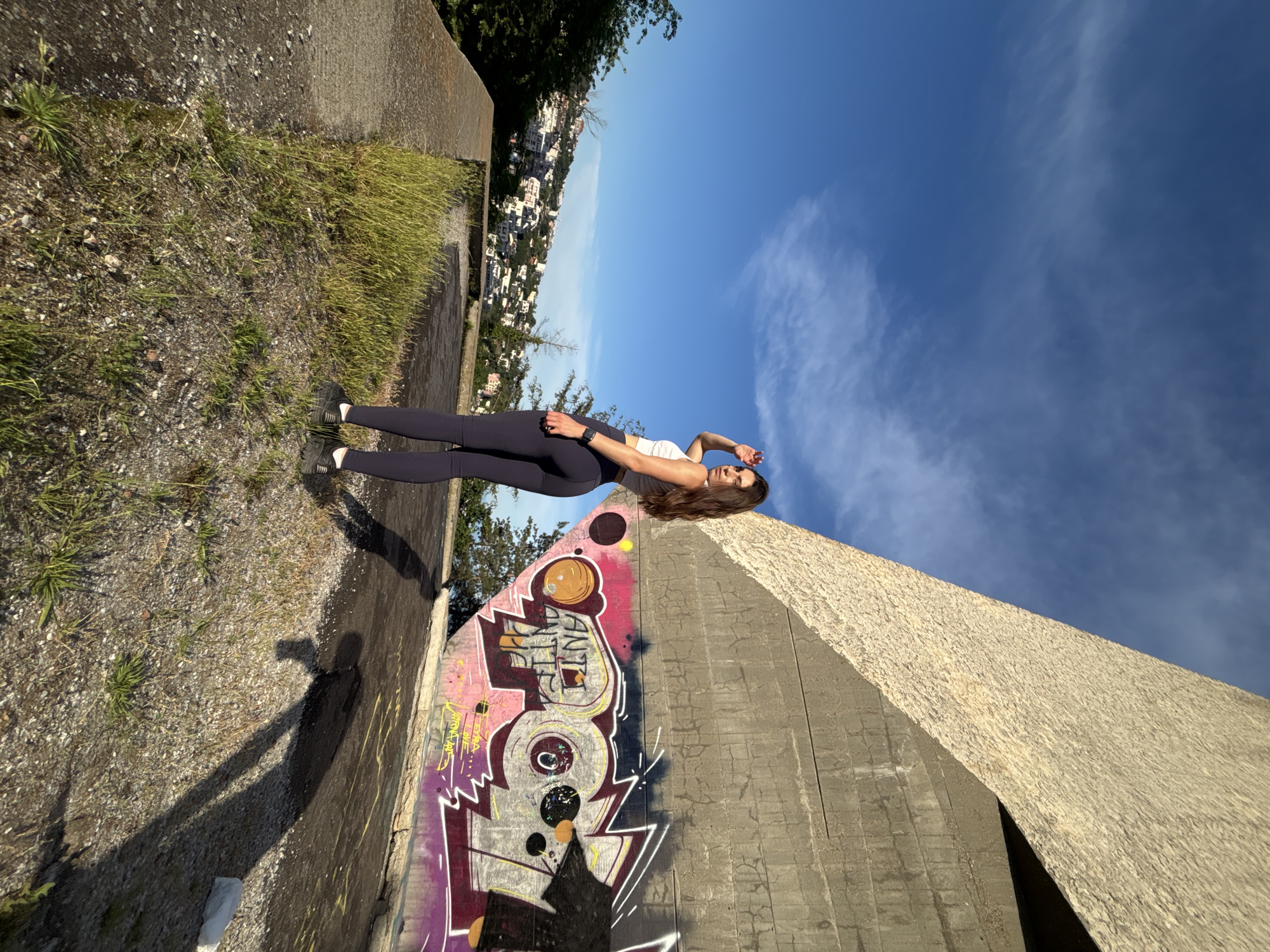 A young woman stands on an elevated concrete platform overlooking the city of Varna, Bulgaria. The woman is facing away from the camera, looking towards the city and shielding her eyes from the sun. She is wearing dark leggings and a white crop top, with athletic shoes. Behind her, a large, angular concrete structure with textured and smooth sections dominates the background. Graffiti covers a section of the concrete wall, featuring bold colors and stylized lettering, including the words "ANTI" and "ANTI THE." The sky is a clear blue with scattered white clouds, and the sun casts long shadows on the ground. The platform is covered in sparse grass and gravel, indicating an outdoor, possibly urban or semi-urban environment. The city of Varna is visible in the distance, with numerous buildings spread across a hillside under the bright sky.