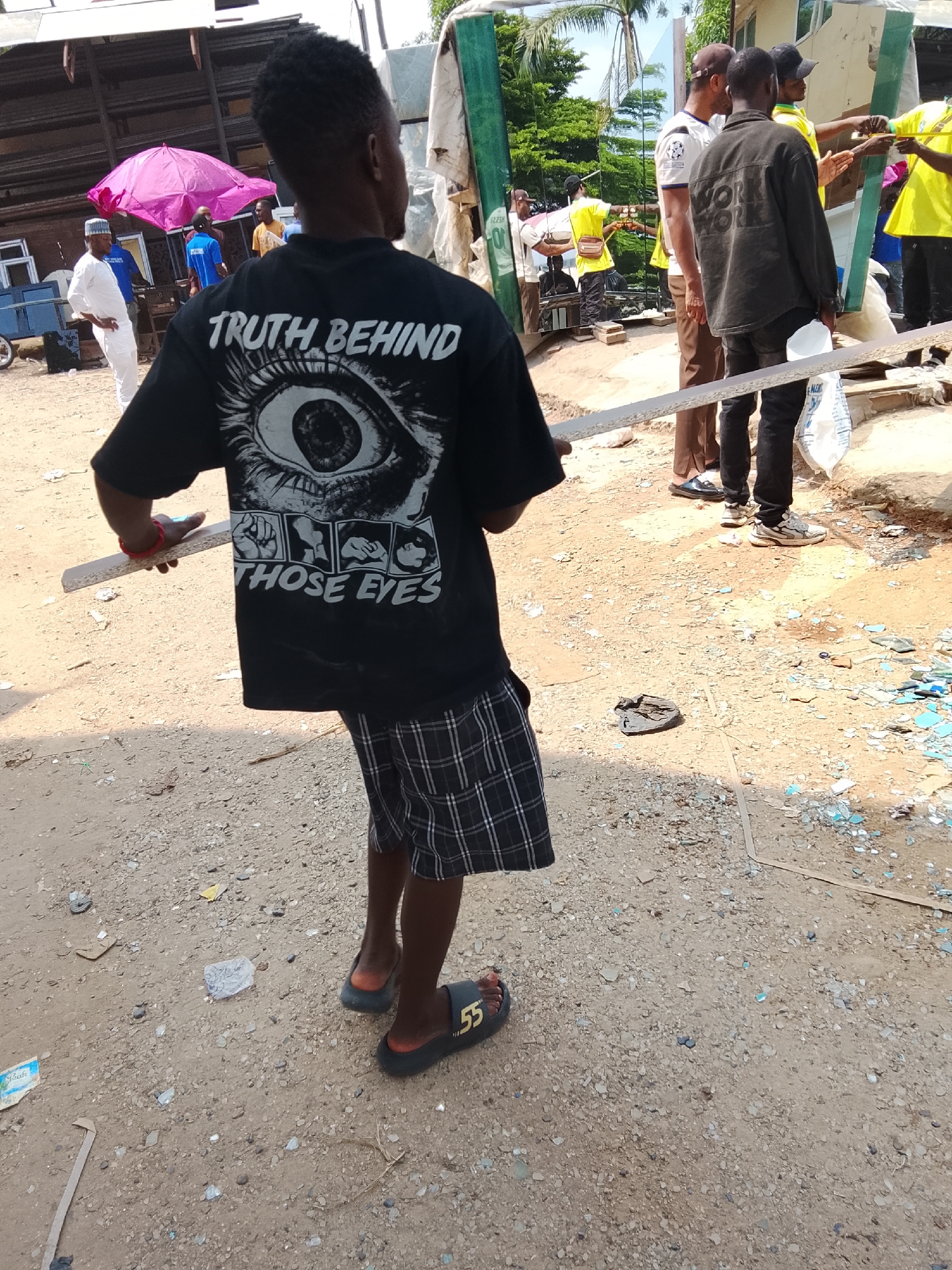 A group of people are gathered in an outdoor area, possibly a construction or demolition site, with scattered debris including glass shards and building materials. In the foreground, a young person wearing a black t-shirt with a graphic design of an eye and the text "TRUTH BEHIND THOSE EYES" and checkered shorts walks away from the camera. The t-shirt's graphic includes several framed images of people's faces. The person is wearing black slide sandals with the number "55" on them. Other individuals in the background are seen wearing various clothing, including a man in a white traditional outfit and cap, another in a yellow shirt and dark pants, and others in casual attire. A pink umbrella is visible in the background, providing shade. The ground is dirt and gravel, with broken glass scattered widely. The environment appears to be an informal setting, potentially in a less developed part of a city like Abuja, Nigeria.