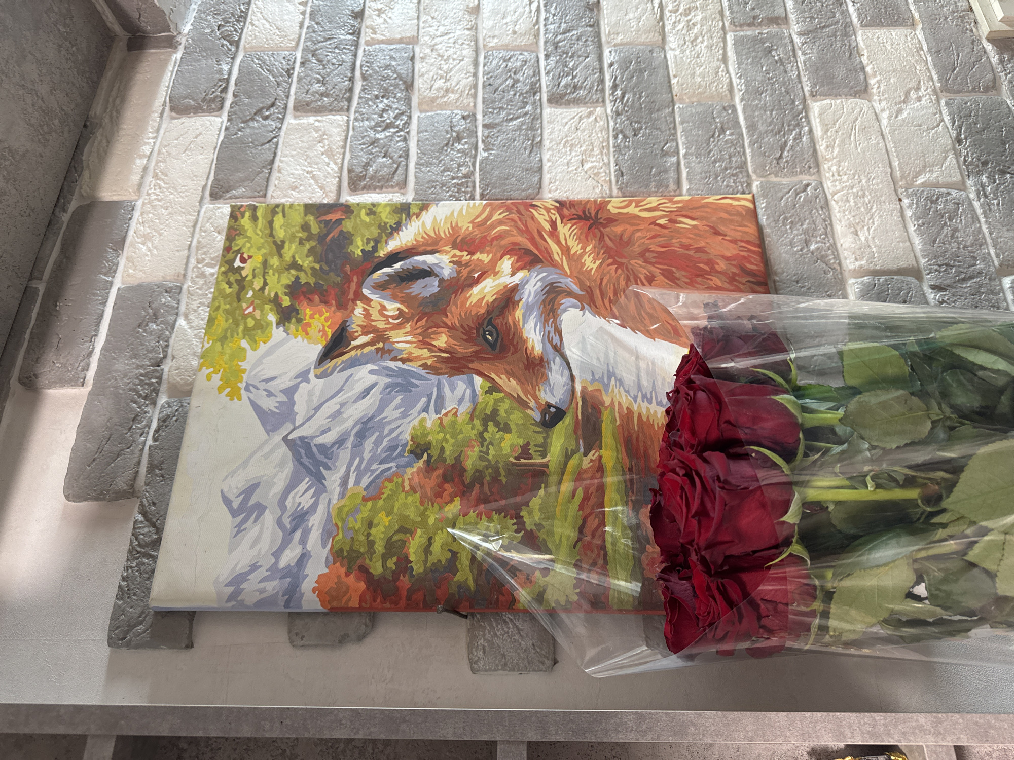 A bouquet of red roses is presented in front of a painting of a fox in a natural landscape. The painting depicts a fox with orange and white fur, facing left, with a backdrop of mountains, trees in autumn colors, and a body of water. The painting is covered in clear plastic wrap. The scene is set against a brick wall with varying shades of gray and white bricks. The location is identified as Chaikovsky, Russia.