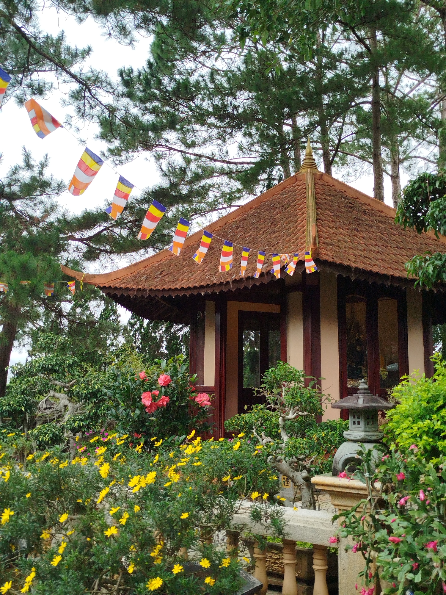 The image depicts a small, ornate temple-like structure with a terracotta tiled roof, adorned with strings of colorful prayer flags. The structure is nestled within a lush garden setting, characterized by abundant greenery, vibrant yellow flowers in the foreground, and a few blooming pink flowers. Several pine trees are visible in the background, reaching towards a cloudy sky. A stone lantern sits on a low wall in the lower right quadrant, adding to the serene ambiance. The architectural style suggests a Buddhist temple. The presence of prayer flags is a common sight in Buddhist religious sites, often found in mountainous or forested areas. This scene is likely located at a temple or pagoda in Da Lat, Vietnam, a city known for its many Buddhist pagodas and its cool, temperate climate which supports abundant floral growth and pine forests. The overall impression is one of tranquility and spiritual contemplation.