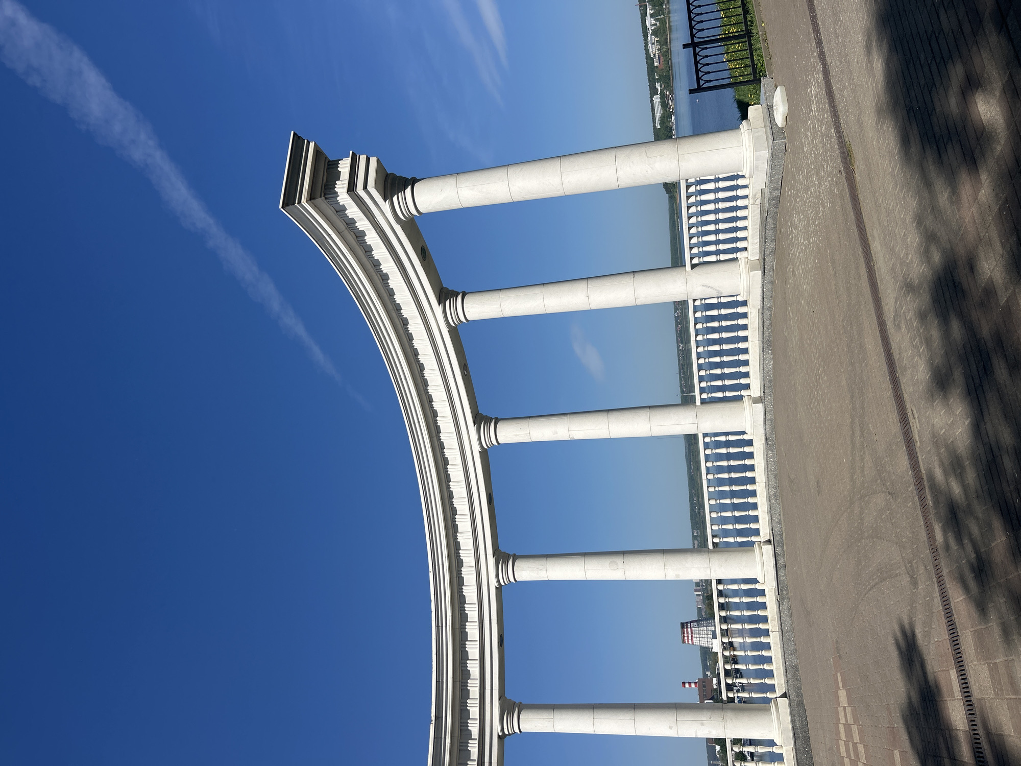 The image depicts a section of the Central Embankment in Izhevsk, Russia. It showcases a curved colonnade made of white stone or concrete, likely part of a decorative architectural structure along the embankment. The colonnade features columns and a balustrade, and it is set against a backdrop of a clear blue sky with a contrail from an airplane. In the background, across the water, are visible buildings and structures of Izhevsk. The foreground shows a paved walkway with shadows of trees falling upon it. The embankment runs along the Izhevsk Pond, a large artificial lake in the city.