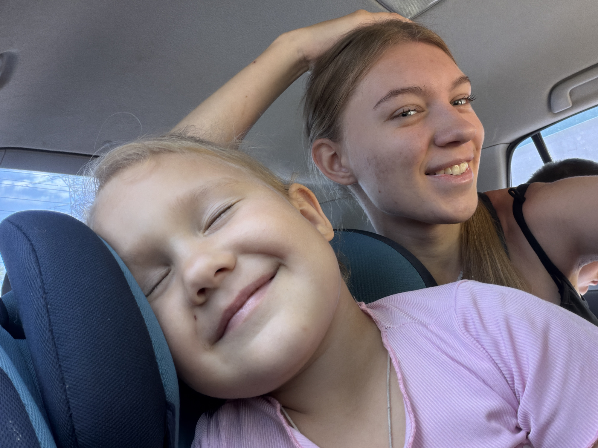In Rostov-on-Don, Russia, two individuals are captured inside a vehicle. A younger girl, with her eyes closed and a gentle smile, is resting her head against a dark blue car seat cushion. She is wearing a pink, ribbed shirt. Beside her, an older girl, with her arm raised and hand resting on her head, is looking towards the camera with a wide smile, revealing her teeth. She is wearing a dark top. The background suggests the interior of a car, with a window visible in the upper right corner showing a glimpse of the sky and possibly a building. The lighting indicates it is daytime.