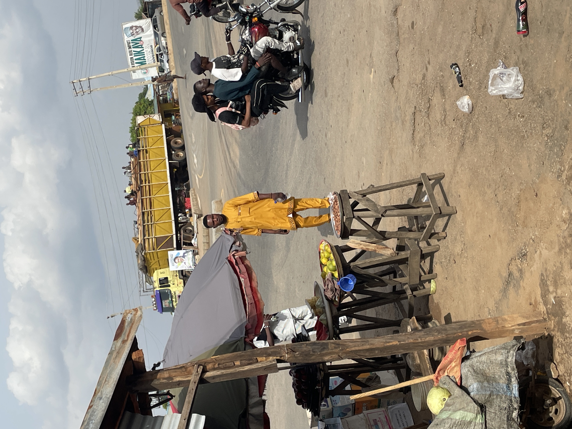 The image captures a bustling street scene in Akwanga, Nigeria. A man in a yellow outfit stands next to a wooden fruit stand with lemons and a blue plastic bowl. An older man is seated under a gray umbrella behind the fruit stand. The front of the stand has items such as watermelons, boxes, and packaging.
Three men are riding on a single motorcycle in the background. A large yellow truck loaded with people is parked on the side of the road. There is a banner on the side of the road. Discarded bottles and plastic bags are on the street. There are telephone poles visible.