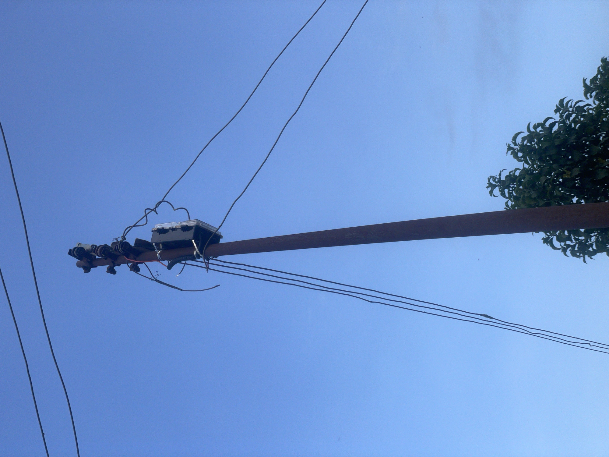 A close-up, low-angle shot captures a utility pole set against a clear, pale blue sky in Udu, Nigeria. The pole, made of rusted metal, is positioned to the right of the frame, with a portion of a leafy green tree visible at the bottom right. At the top of the pole, an electrical transformer box is mounted, surrounded by a tangle of wires and insulators. Several dark, thick electrical lines extend horizontally across the sky from the pole. The scene appears to be outdoors and depicts part of the electrical infrastructure common in urban areas.