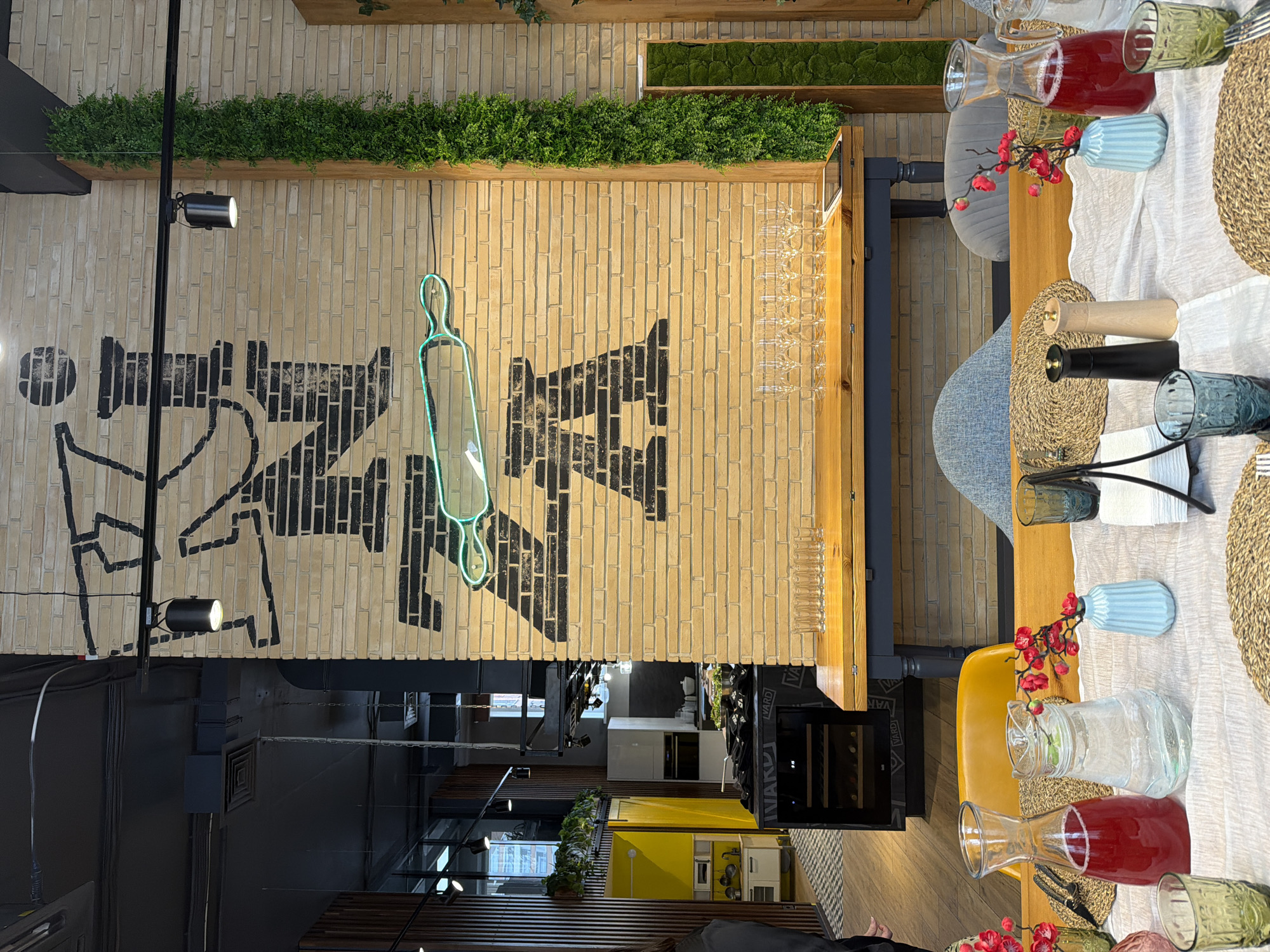 This indoor scene shows a long table set for a meal, positioned in what appears to be a dining area or restaurant. The table is covered with a white cloth and features placemats, glassware, and carafes of beverages. Chairs of varying colors are placed around the table. Overhead, the brick wall is adorned with a mural of a stylized kitchen and a decorative neon rolling pin. Green plants are arranged on shelves along the top of the wall. The view extends into a brightly lit kitchen and other dining spaces. Based on the "AZZA" mural on the wall, the location is likely a restaurant or culinary establishment in Izhevsk, Russia. No people are definitively visible in the image, though there is a hint of a person at the bottom edge of the photo.
