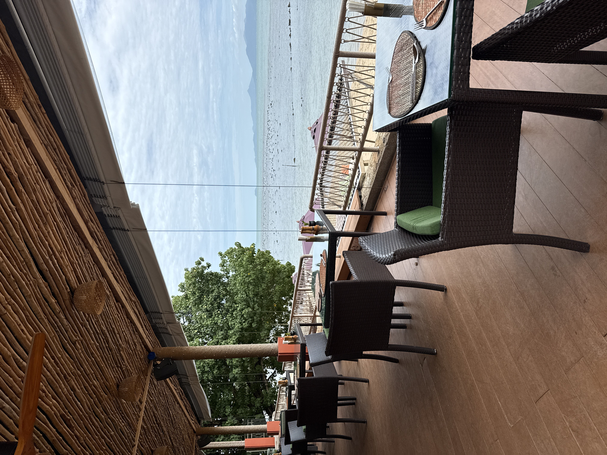 The image shows an outdoor dining area of a restaurant overlooking the ocean in Ko Samui District, Thailand. The area features dark wicker chairs and glass-top tables set on a wooden deck. The ceiling is made of bamboo. The ocean view is partially obstructed by glass windows and a railing. In the distance, several islands are visible under a cloudy sky. The scene is devoid of people.