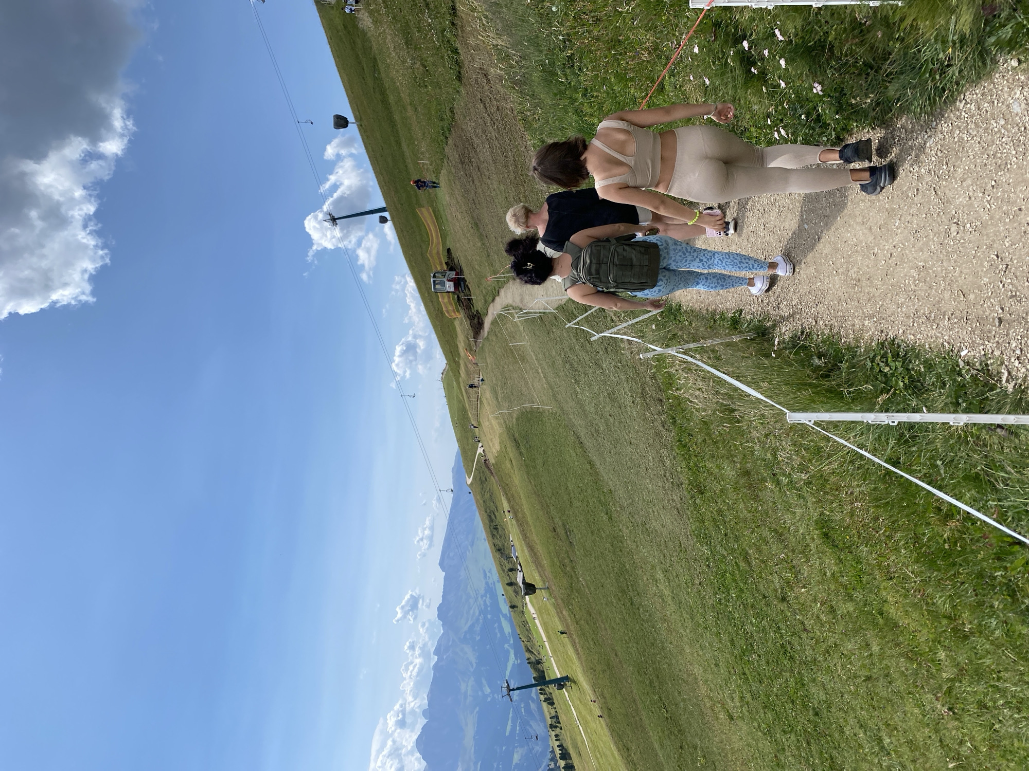 A group of four women walks along a gravel path on a grassy mountainside under a bright blue sky with scattered clouds. The path curves up the slope, bordered by white posts and thin white cord fencing. The women are dressed in athletic wear, with one in a khaki-colored matching top and leggings, another in patterned blue leggings and a green backpack, and the other two in casual tops and shorts. In the background, ski lifts are visible ascending the steep grassy terrain, and in the far distance, a range of jagged mountains creates a scenic backdrop. The area appears to be a hiking trail or a mountain resort.