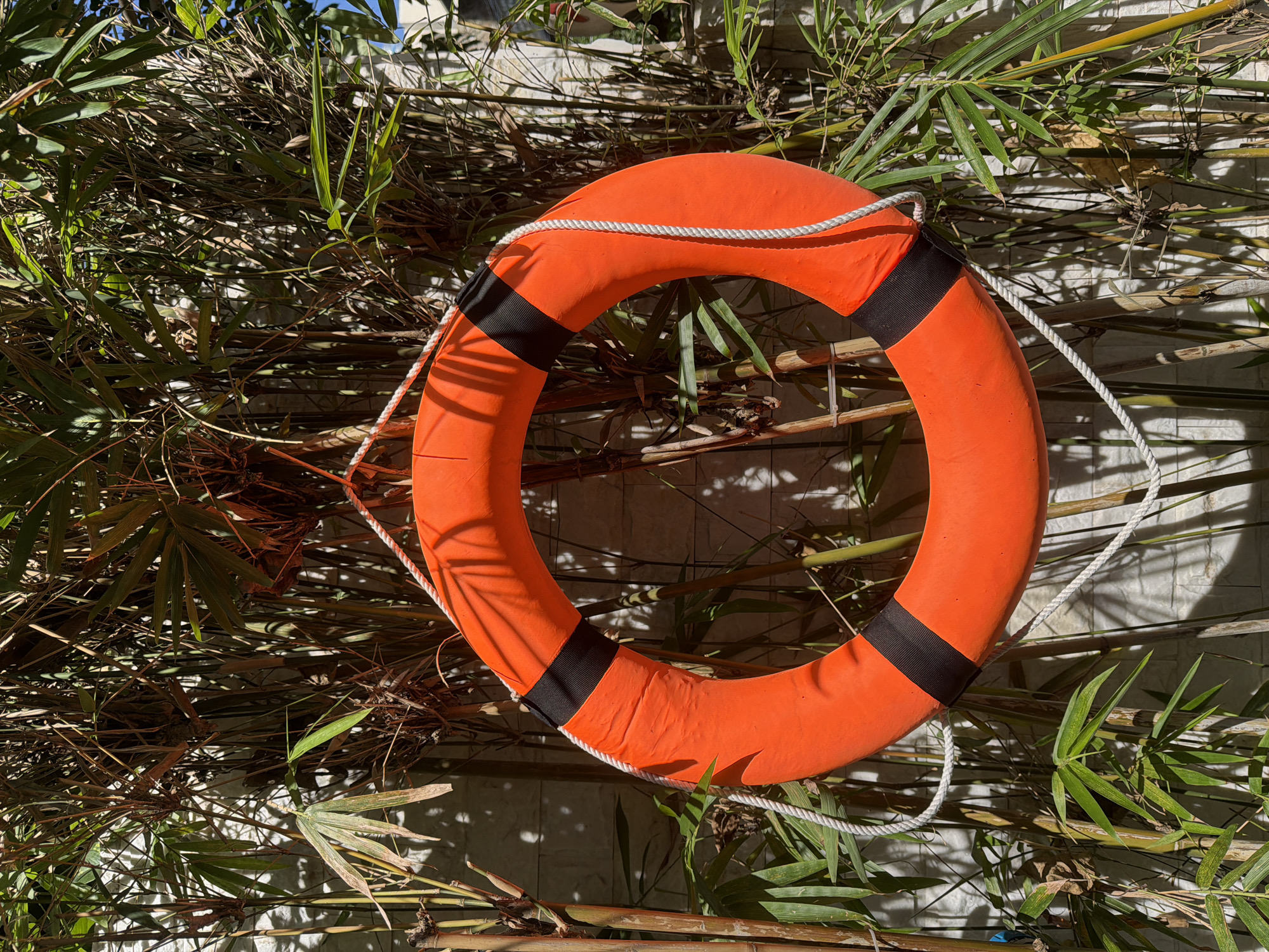 An orange life preserver with black bands and a white rope is suspended in front of a wall covered with bamboo stalks and leaves. The wall appears to be made of light-colored, textured bricks. Sunlight casts shadows from the bamboo onto the wall and the life preserver. The scene suggests an outdoor environment, possibly in a garden or poolside area. No people are present. The image is taken from a slightly low angle, looking up at the life preserver. The location is identified as Nha Trang Ward, Vietnam.