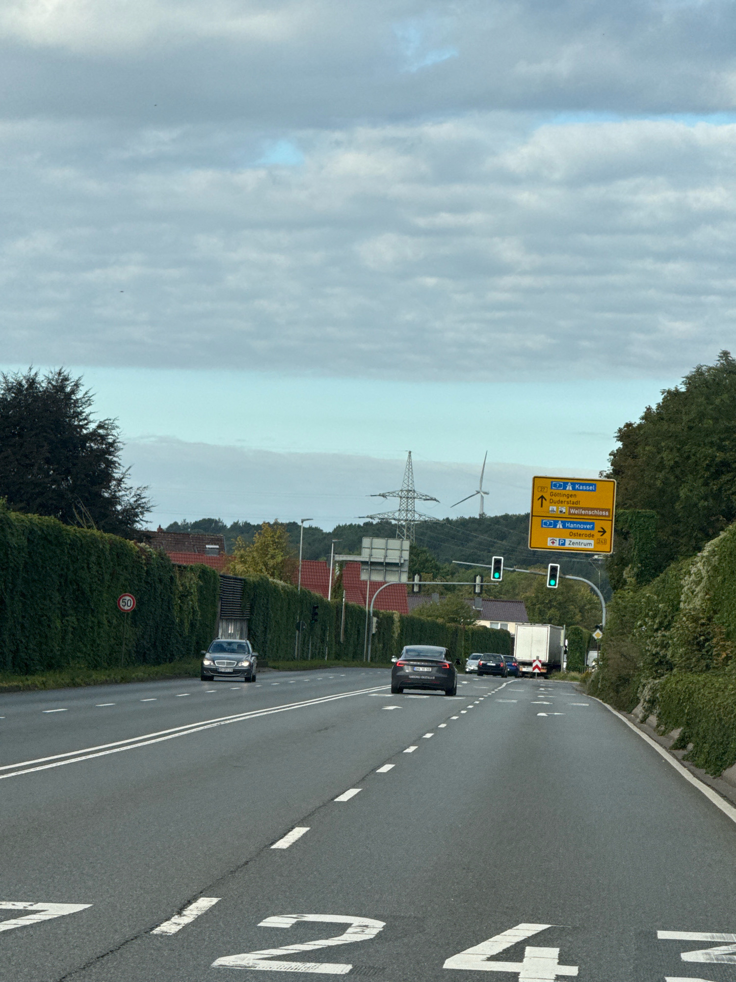 The image depicts a view from a vehicle traveling on a two-lane road in Herzberg, Germany. The road is asphalt with white lane markings and large white numbers painted on it, indicating lanes or distances, with '12' visible on the left lane and '43' visible on the right lane. The surroundings are characterized by a dense green hedge lining the left side of the road, with a circular speed limit sign displaying '50' mounted on it. On the right side of the road, there is also dense green foliage.
In the distance, there are buildings with red-tiled roofs and trees. Overhead, the sky is cloudy with patches of blue. A significant element in the background is a tall electrical pylon and a wind turbine, suggesting a rural or semi-rural setting.
Traffic on the road includes several vehicles. In the foreground, there is a dark-colored sedan in the left lane, followed by other cars. Further ahead, in the right lane, a dark-colored sedan is visible, and behind it, a white truck with a red and white striped rear is present. Traffic lights are visible at an intersection, with green lights illuminated.
A yellow directional signpost is prominently displayed. It indicates directions to "Kassel" via the A7, "Göttingen," "Duderstadt," and "Wellenschloss" for the left lane. The right lane is directed towards "Hannover" via the A7 and "Osterode," with "Zentrum" (center) also indicated. The road numbers "27" and "243" are also present on the sign. The scene captures a moment of travel on a well-maintained road under an overcast sky.