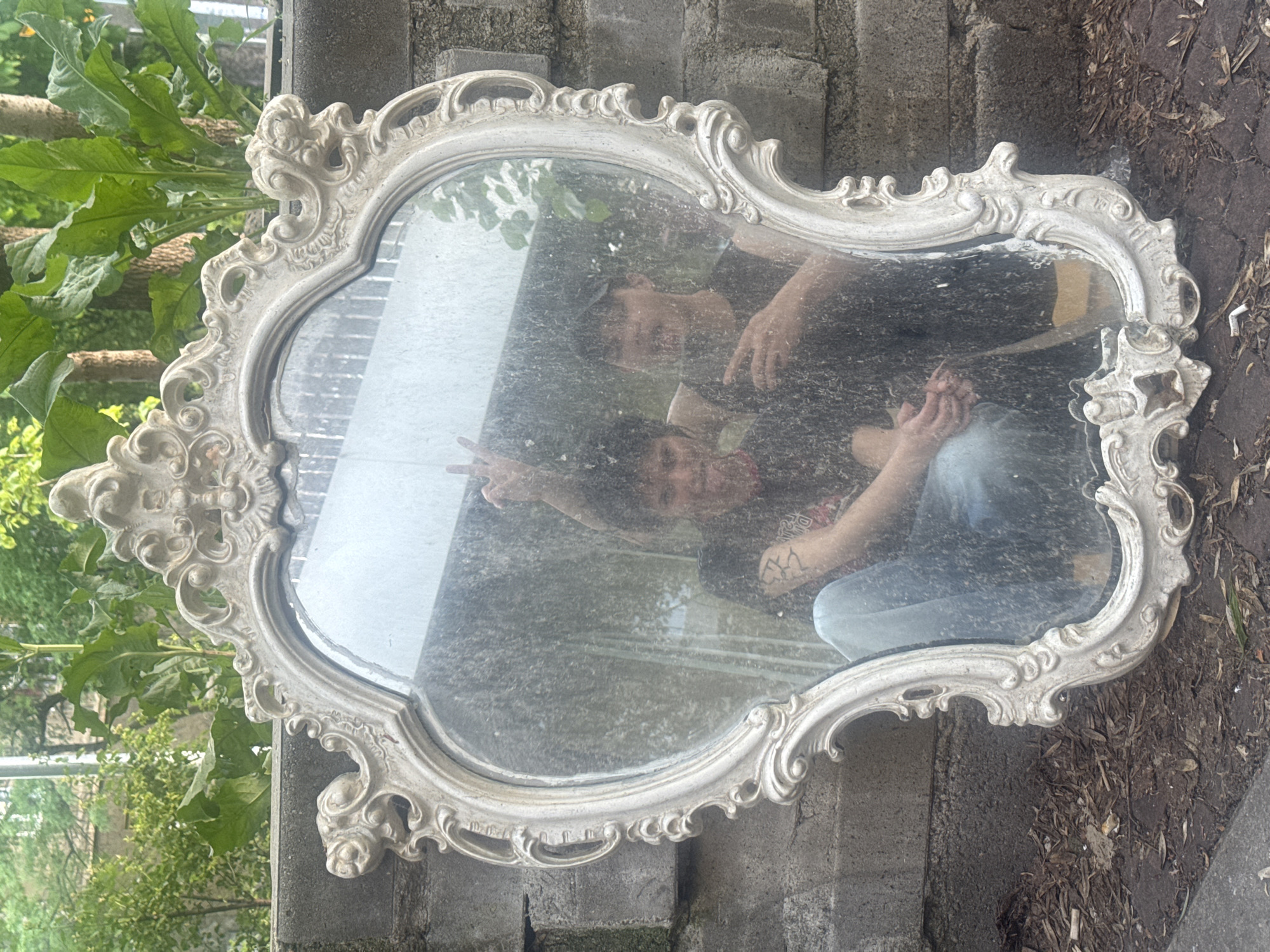 The image shows an ornate, antique-style mirror resting against a low brick wall. The mirror's frame is white with decorative swirls and flourishes. The mirror is reflecting two people, one flashing a peace sign and the other with their arm around them. One person has a tattoo on their arm. The mirror itself is dusty. The background consists of green foliage on the left and dry leaves and brick on the right. The exact location cannot be determined from the image, but it is mentioned to be in the city of Sofia, Bulgaria.