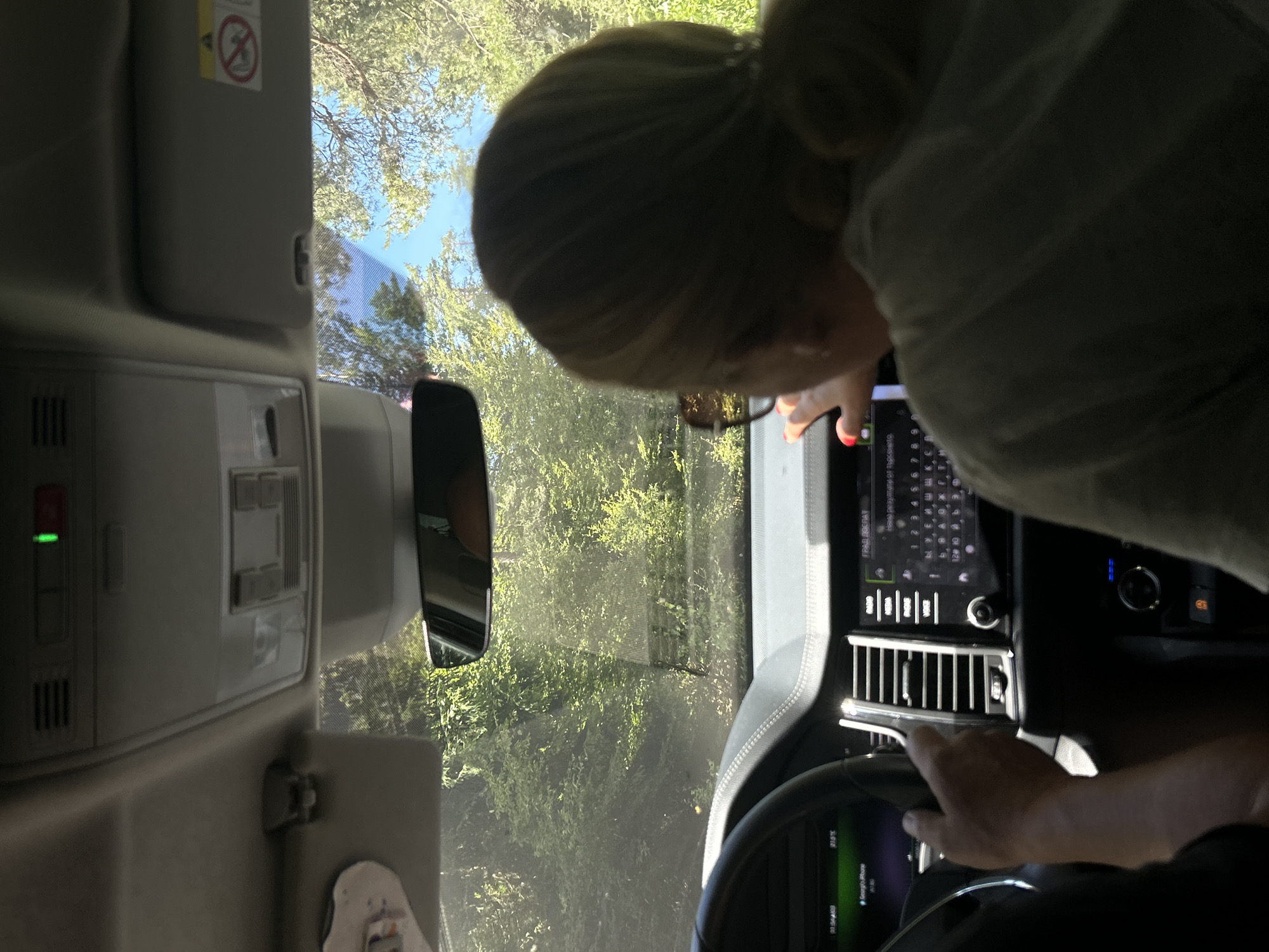 The image is an interior shot of a car, taken from the back seat, looking towards the front. A person's hands are visible on the steering wheel, and another person's back and head, with blonde hair pulled into a ponytail and wearing sunglasses, is in the passenger seat, interacting with the car's touchscreen display. The car is driving on a road surrounded by lush green trees and foliage, suggesting a rural or natural setting. The sky is visible through the windshield and trees, appearing blue with some branches. The car's interior shows a dashboard with a navigation screen displaying a keyboard layout, a steering wheel, and air vents. The context suggests the car is currently in motion, driving through a scenic route possibly near Batak, Bulgaria, given the information provided. The overall impression is a snapshot of a road trip.