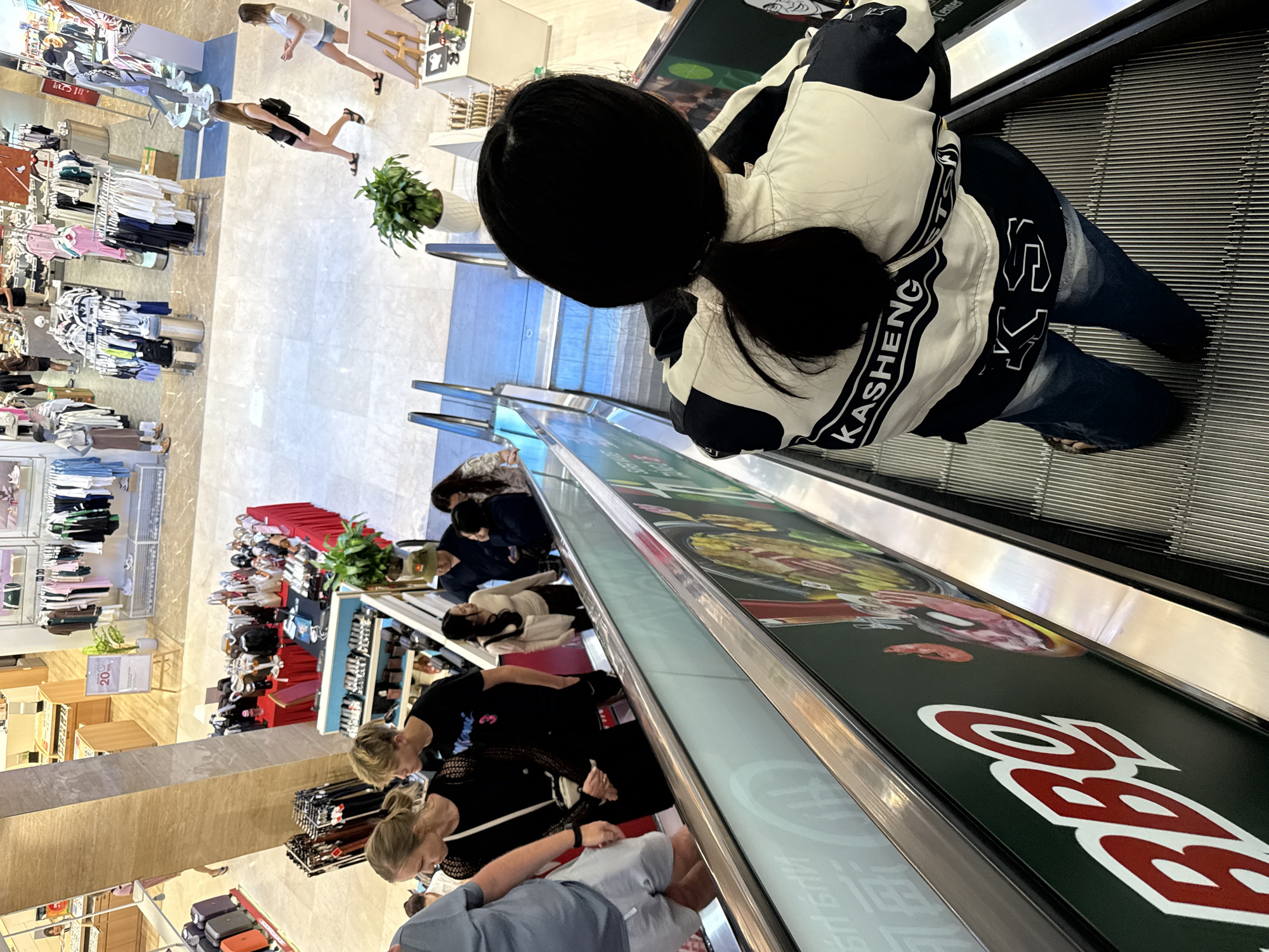 In a brightly lit shopping mall in Nha Trang Ward, Vietnam, people are seen navigating escalators and browsing retail displays. The scene depicts a typical day of commerce and leisure.
Several individuals are the primary subjects. One person, viewed from behind, is on an escalator, wearing a black and white jacket with the text "KASHENG" visible. Another group of people is gathered on a lower level, appearing to shop or observe merchandise. This group includes individuals with blonde and dark hair, dressed in casual attire such as t-shirts and shorts. Further down, other shoppers are visible, some looking at racks of clothing.
The setting is a modern shopping mall with polished floors and well-stocked retail shelves. Displays of clothing, accessories, and possibly food items are evident. The escalators are prominent features of the scene, indicating movement between different levels of the mall. The overall atmosphere seems relaxed and functional, characteristic of a commercial environment.
The activity is primarily shopping and moving through the mall. People are engaged in browsing goods, walking, and using the escalators. There are no overt indications of a specific event or urgent activity.
Notable details include the clear, bright lighting typical of indoor retail spaces. The presence of plants suggests an attempt to enhance the mall's aesthetic. The visible text on the person's jacket and the large, stylized red text on the side of the escalator, which appears to be part of an advertisement, add context to the commercial nature of the location. The clothing worn by the individuals suggests it is daytime and likely warm weather outside, consistent with a tropical location like Nha Trang. The general demeanor of the people suggests they are engaged in everyday activities.