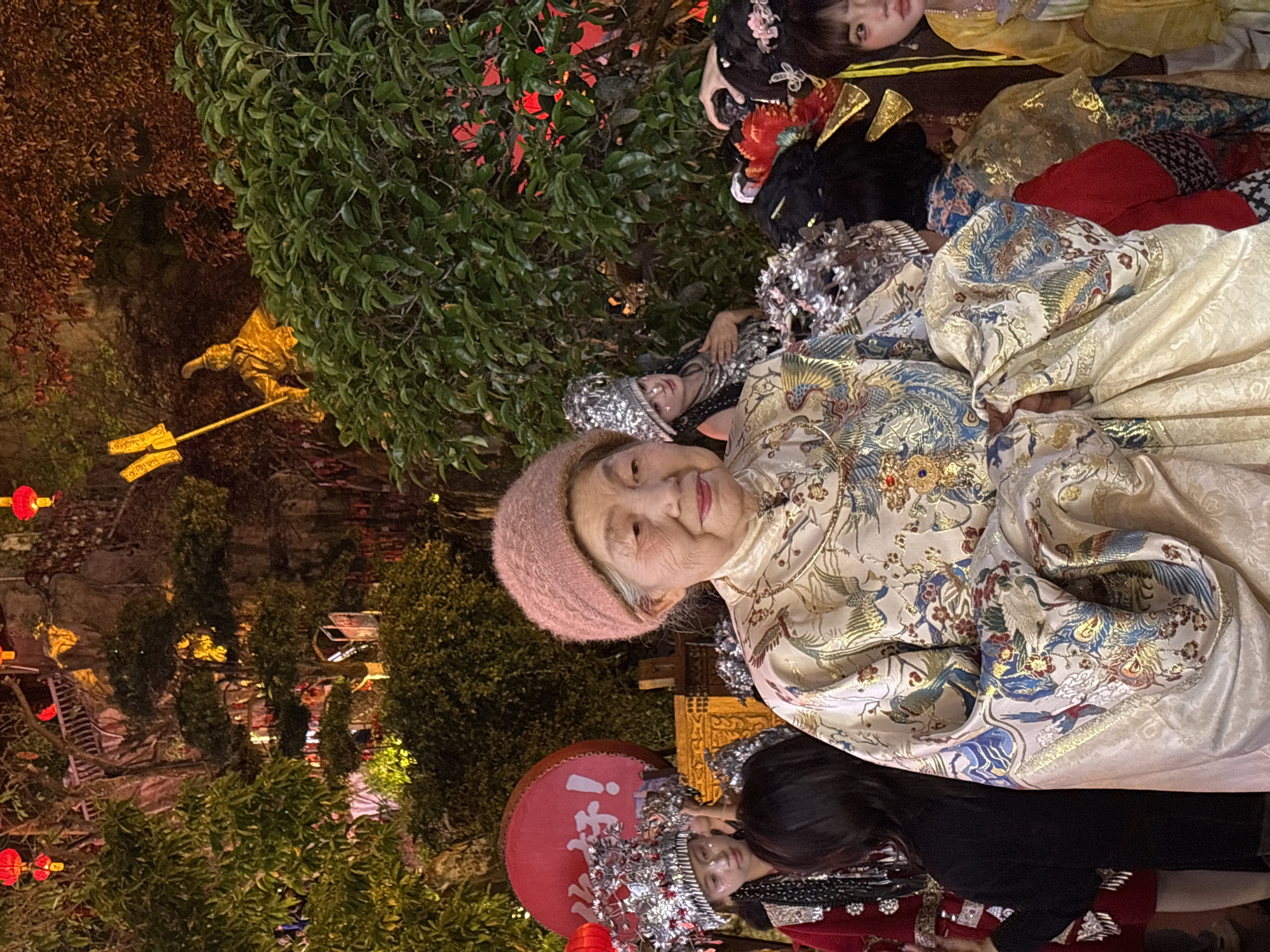 In a low-angle, nighttime shot, a group of people in traditional Chinese attire gathers in what appears to be a scenic area. The central subject is an elderly woman with a pink knit cap, wearing a patterned cream and blue robe adorned with bird motifs. She is positioned to the right of the frame, looking towards the camera with a slight smile. To her left and below, another individual is seen wearing an elaborate silver headpiece and a black top with red accents, holding a red circular object with Chinese characters.
Behind the elderly woman, several others are seen in similar traditional clothing, some with ornate headwear. The composition is tight, with the figures closely positioned together. The background features lush greenery, lit with red lanterns, creating a warm and festive atmosphere. Golden decorative figures are partially visible among the trees. Based on the presence of traditional Chinese architecture and the lanterns, the location appears to be within the Yuzhong District of China.
