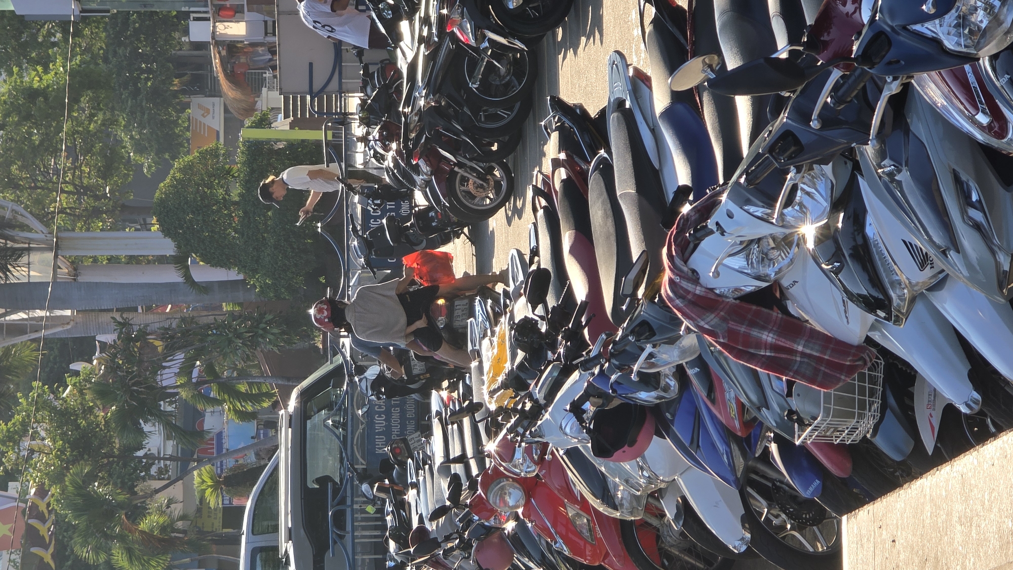 The image captures a street scene in Nha Trang, Vietnam. A dense row of parked motorbikes occupies the foreground, showcasing various makes and models, with Honda and Yamaha logos visible. A man in a gray shirt and helmet straddles one of the motorbikes, appearing to interact with it. Another man in a white shirt and cap is standing in front of other motorbikes in the distance, looking at a phone. In the background, a street sign is partially visible. The environment includes trees and buildings.