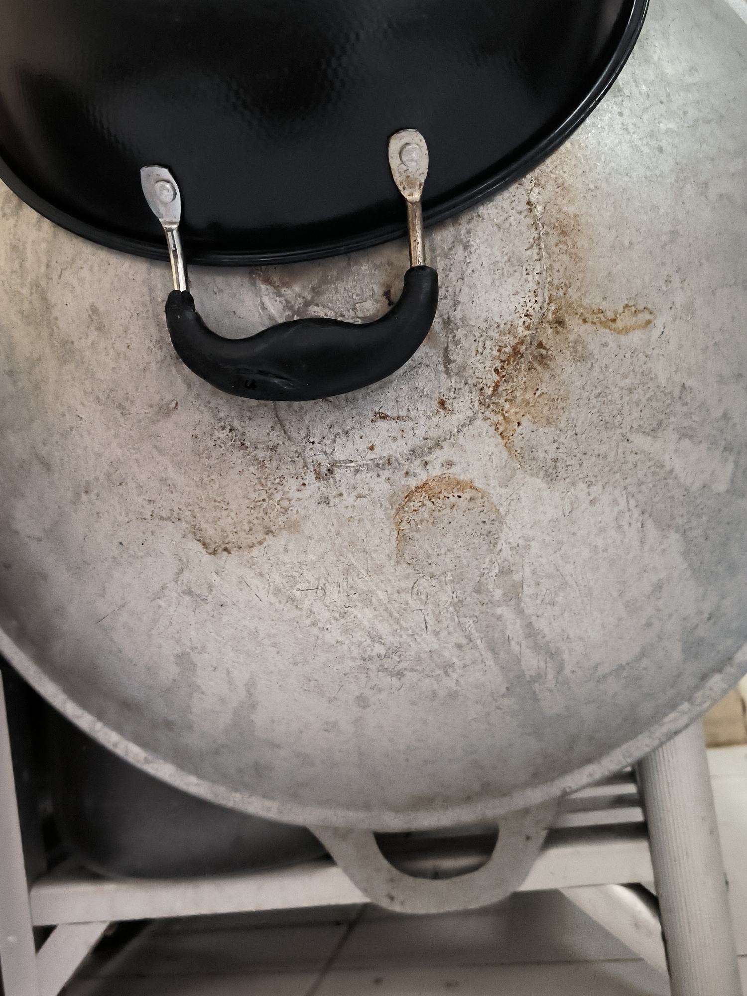 A large, well-used metallic pot or wok with a matte black lid is depicted from an overhead perspective. The pot's main body is made of a light-colored, likely aluminum, material, exhibiting numerous scuffs, scratches, and patches of yellowish-brown residue, consistent with cooked-on grease or food stains. Two distinct circular, stained areas are visible on the pot's surface.
The black lid features a textured, possibly hammered or dimpled, surface. A black, U-shaped handle, likely made of plastic or rubber, is centrally mounted on the lid. This handle is secured by two metallic, silver-colored fasteners, each appearing to be a riveted tab.
The pot rests on a light-colored, possibly metallic, stand or frame, with part of a dark, indeterminate object visible beneath the pot. A section of a light-colored tiled floor is discernible in the lower background. The overall scene suggests a kitchen or cooking area.