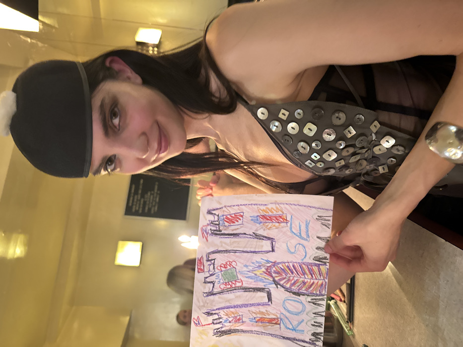 A woman wearing a black hat and a gray top adorned with square buttons holds up a child's drawing. The drawing features colorful crayon scribbles of what appears to be a castle, a feather, and the names "ROSE" and "KOKO." Light fixtures and a dark plaque are visible in the background, along with blurred figures reflected in a mirror. The woman is also wearing a silver bracelet on her right wrist.