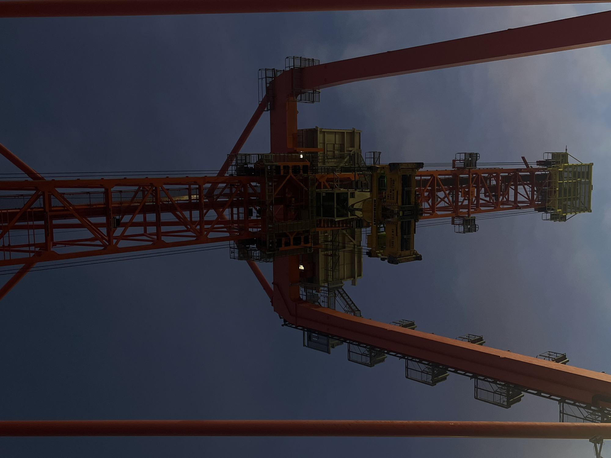 The image shows a large orange industrial crane structure against a blue sky. The crane is made of a complex network of metal beams and platforms. The location is Unknown, Unknown. There are no people visible.
