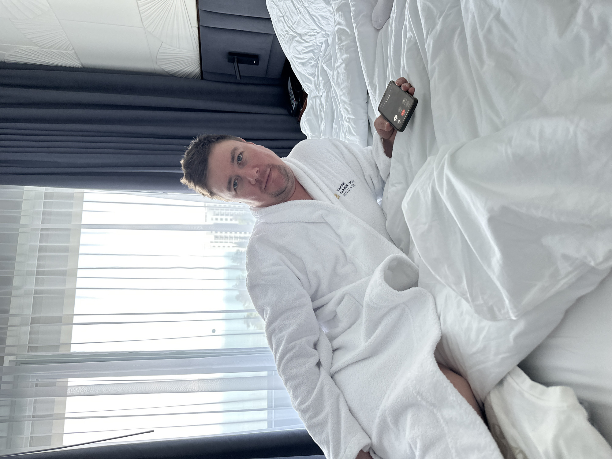 This media file captures a man relaxing in a hotel bed, appearing to be interrupted by a phone call. The man, who is middle-aged with short, dark hair, is lying on his left side, propped against pillows with white sheets. He looks directly at the camera with a neutral expression. He is wearing a white terry cloth bathrobe.
The bathrobe features embroidered text on its left chest area, which clearly reads "MARRIOTT SOCHI KRASNAYA POLYANA HOTEL," confirming the location as Sochi, Russia. In his left hand, the man holds a black smartphone, its screen illuminated with an incoming call interface. The caller's name "KHARAN" is visible at the top, along with the standard "Decline" (red button) and "Accept" (green button) options.
The setting is a well-lit hotel room. To the man's left, a large window is visible, covered with sheer white blinds or curtains that allow bright daylight to stream in, suggesting it is daytime. Out of focus buildings can be seen outside the window. Behind the bed, a dark grey upholstered headboard or wall panel is present. The ceiling, partially visible at the top of the frame, has a decorative white textured pattern resembling a fan or shell design. The overall scene suggests a moment of leisure in a comfortable hotel environment.