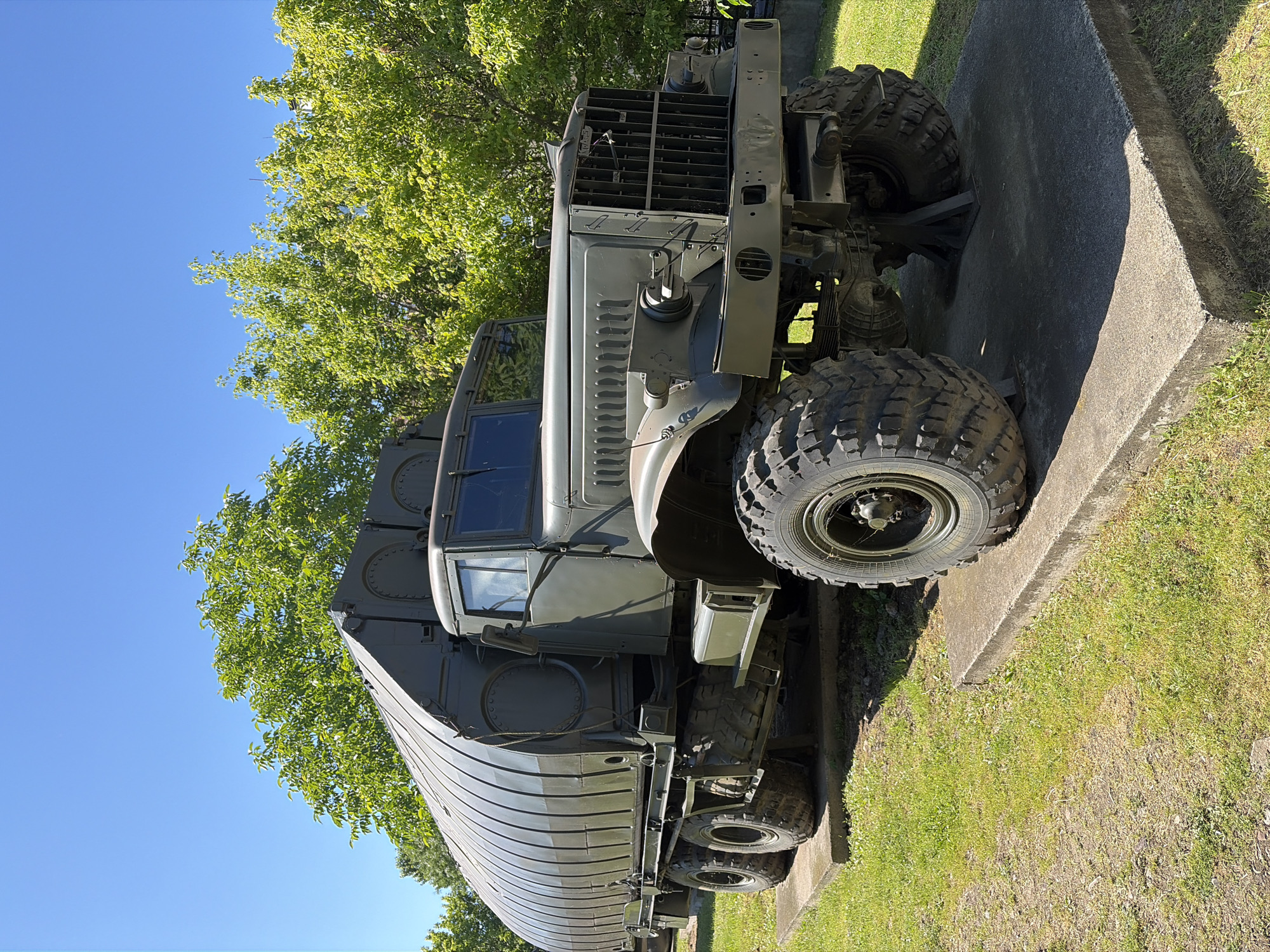 A large, olive-drab, military truck with a rounded cargo area is parked on a concrete pad on a grassy lawn. The truck has large, rugged tires. In the background, lush green trees are visible against a clear blue sky. The truck is a KRAZ-214B, a Soviet-era heavy off-road truck. This specific truck is likely on display at the National Museum of Military History in Sofia, Bulgaria, a common location for such vehicles. There are no people visible in the image. The truck is stationary and appears to be in good condition.