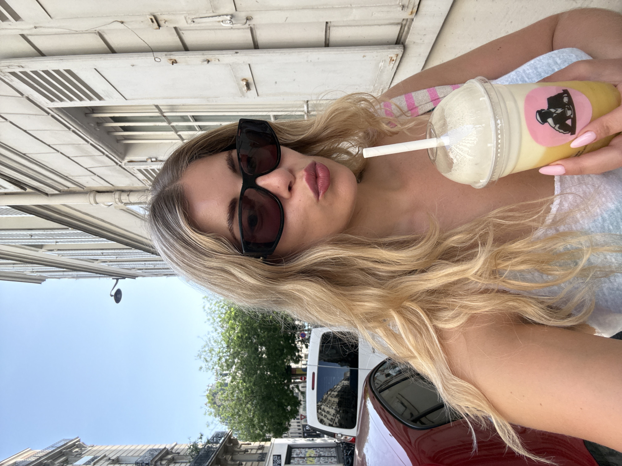 A medium shot shows a fair-skinned person with long blonde hair, wearing sunglasses, and holding a yellow-and-white smoothie with a pink sticker in a clear plastic cup with a straw. The person is positioned so that the background includes a white building with closed shutters and a street scene with trees and a white van. The buildings suggest the location is Paris, France.