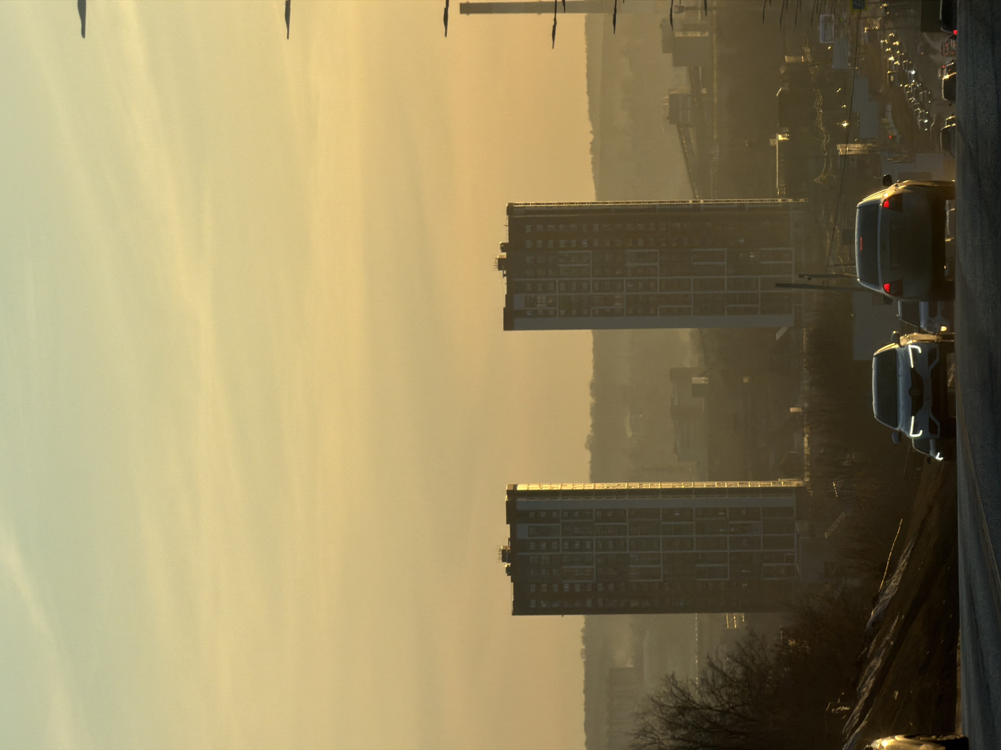 The image shows an urban scene in Izhevsk, Russia at dusk. Two high-rise residential buildings stand prominently in the middle distance, with symmetrical rectangular structures and numerous windows. A highway runs along the right side of the image, with vehicles, including cars and vans, visible. The background landscape is hazy and indistinct, with hints of more buildings and possibly a river valley. The sky is a yellowish-golden color, suggesting either sunrise or sunset. Bare tree branches are visible in the foreground. There is a street light on the right near the foreground. There are what look to be power lines stretching into the background.