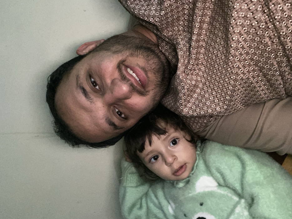 A man and a child lie together facing the camera. The man has dark hair, a beard, and wears a patterned brown shirt. His head is tilted. The child, nestled close, wears a light green sweater with a bear design. Both have fair skin. The background appears to be a plain, light-colored surface. The perspective is slightly rotated, creating a casual, intimate portrait.