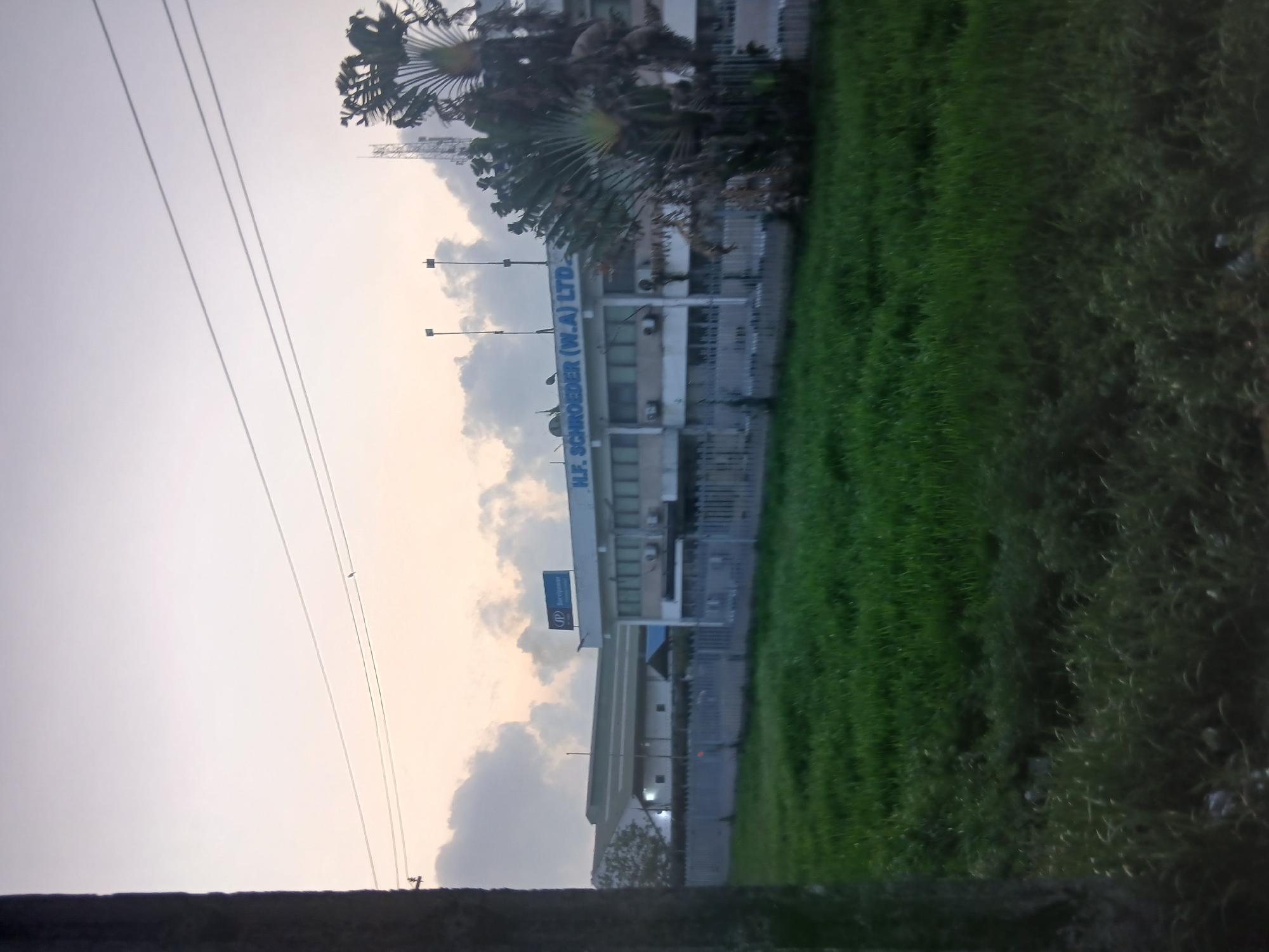 The image shows a building with the sign "H.F. SCHROEDER (W.A.) LTD." in Lagos, Nigeria. The building is light-colored and multi-storied. There is a grassy area to the right of the building. Overhead power lines are visible in the sky. A tree is adjacent to the building, and clouds are in the sky. A sign with the SP logo is also visible on the building.