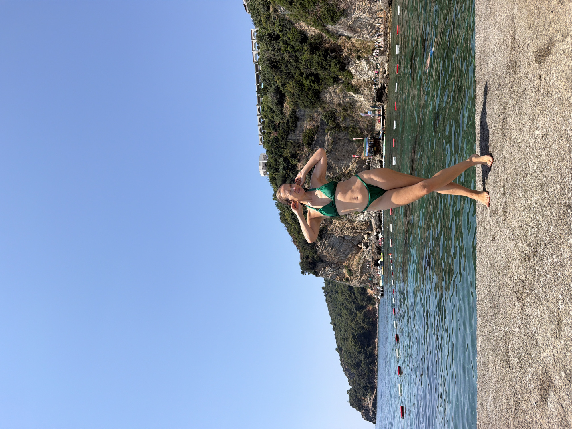 In Budva, Montenegro, a woman stands on a concrete pier on a sunny day, posing for a photo in a green bikini. She has her hands on her head and her legs crossed. The water is clear and blue, with a rope and buoys marking a swimming area. In the background, a rocky cliff covered in green trees rises from the sea. Buildings with arched windows are visible on top of the cliff. People can be seen on the beach and in the water.