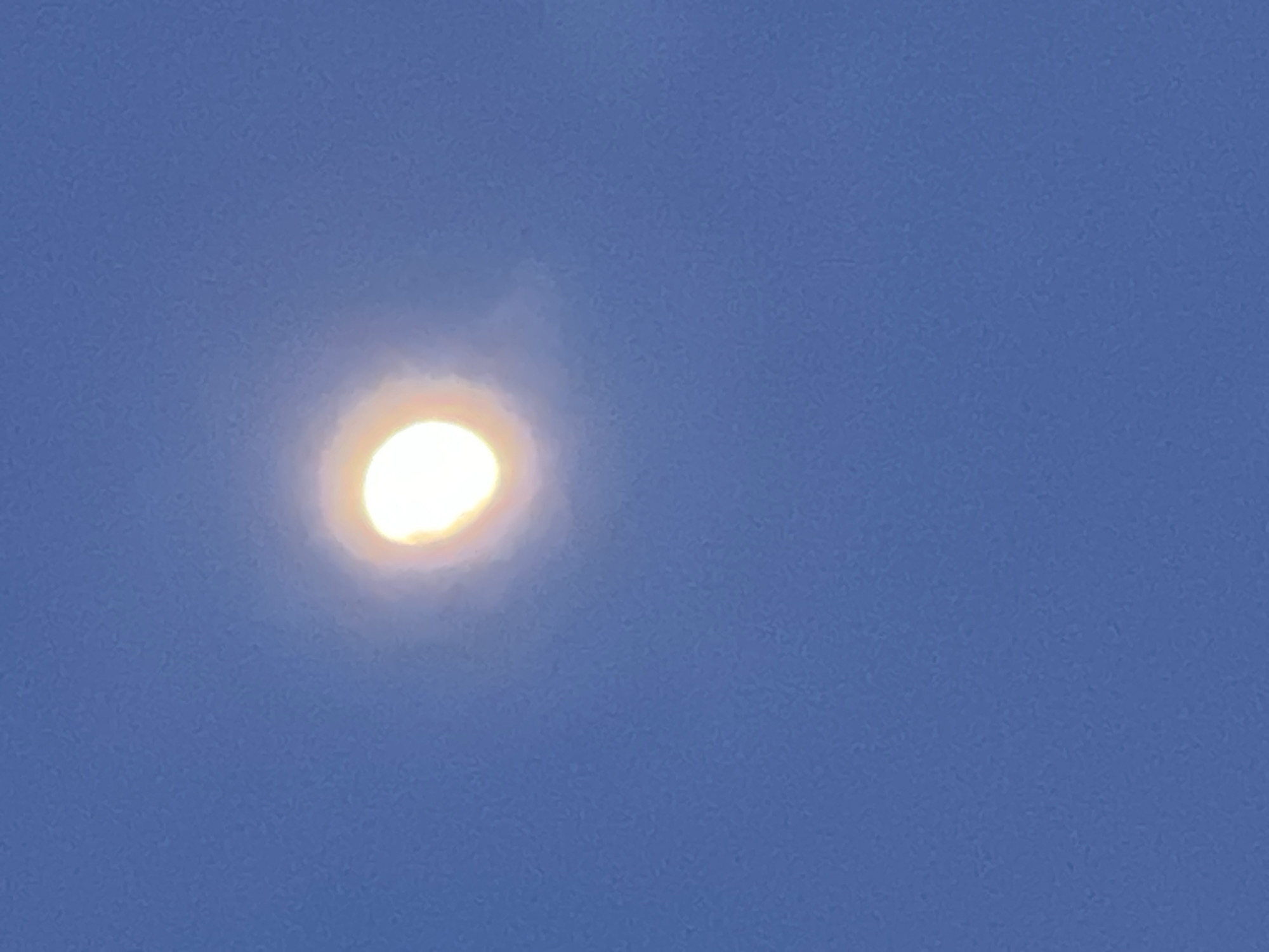 The image displays a clear blue sky with a prominent, intensely bright, and glowing celestial body, likely the sun, positioned slightly to the left of the frame's center. The object appears as an overexposed, roughly elliptical shape, radiating a strong white-yellow light, with a soft, diffused halo or atmospheric glow surrounding it. The sky is a consistent medium-blue color, indicating daylight conditions. There are no clouds visible in the wider sky, though the halo around the light source suggests a thin haze or atmospheric diffusion. No other subjects, objects, or activities are depicted. There is no visible text. The scene is set outdoors in Chaikovsky, Russia, during what appears to be a clear day, possibly early morning or late afternoon given the quality of light.